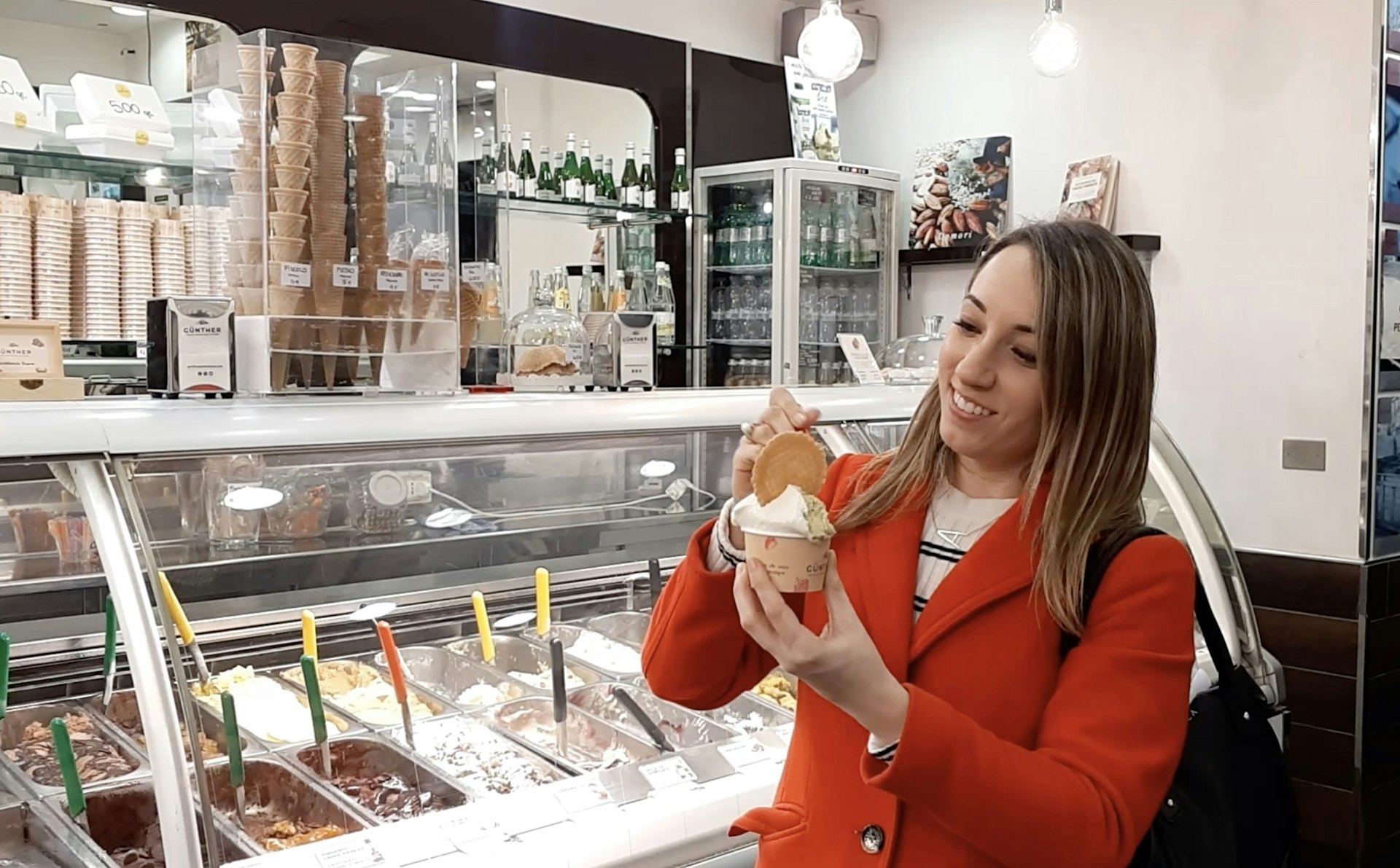 Wearing a red jacket and smiling, Alexandra holds a cup of gelato in the shop