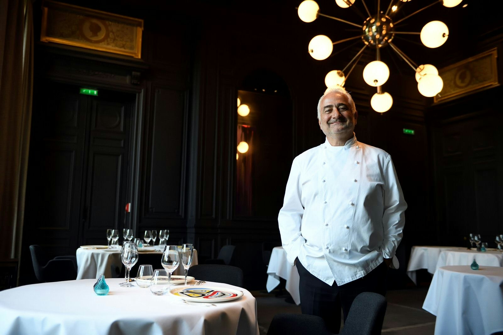 French chef Guy Savoy smiles at the camera wearing his chef's jacket in the dining room of his Michelin three-starred restaurant Guy Savoy de Paris in the Monnaie de Paris. The dark dining room is illuminated by an unusual, globular light fixture. 