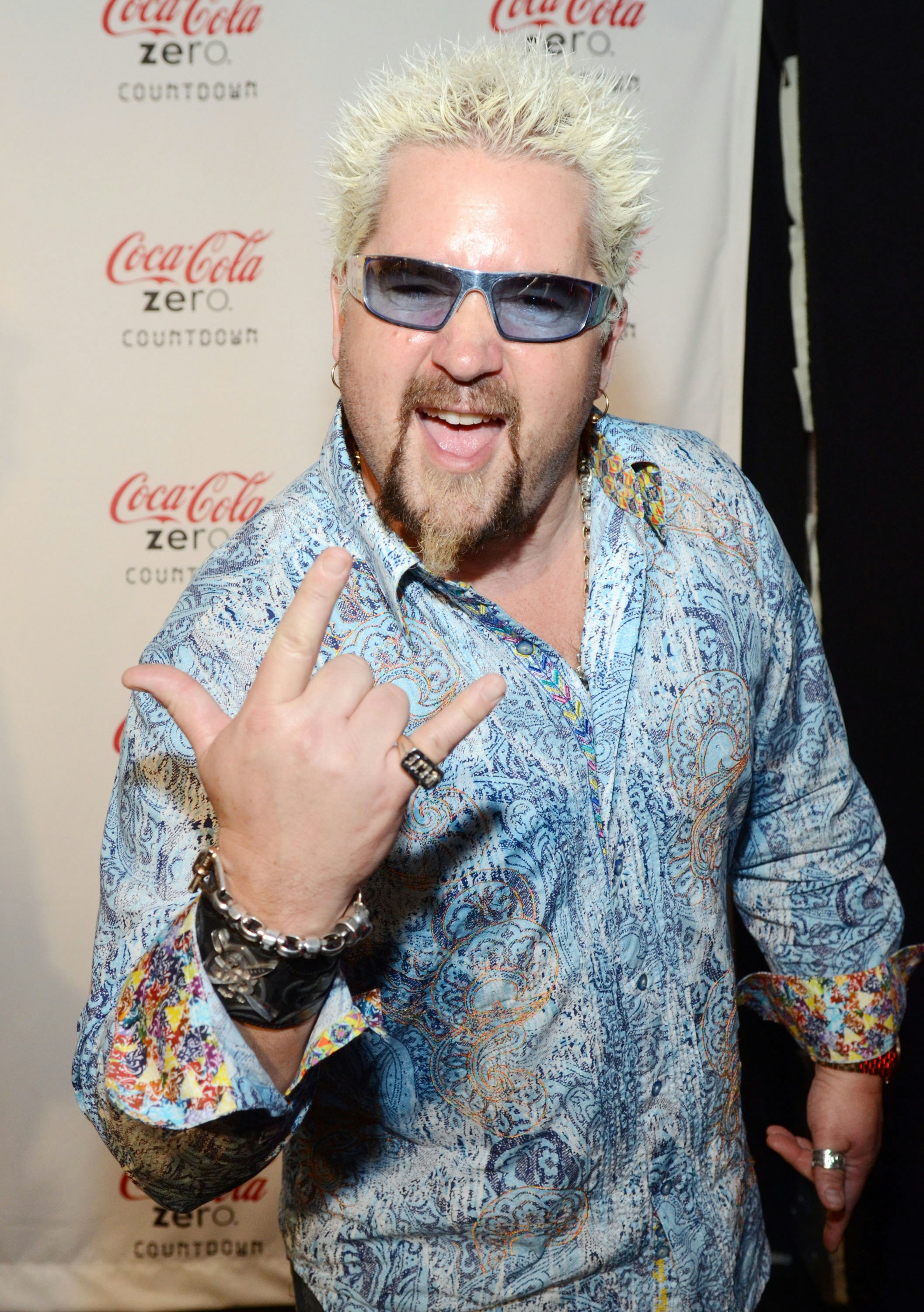 Celebrity chef Guy Fieri, dressed in a light blue paisley button-up shirt with light blue shades, throws up a hand signal to the camera; Columbus vs Ann Arbor