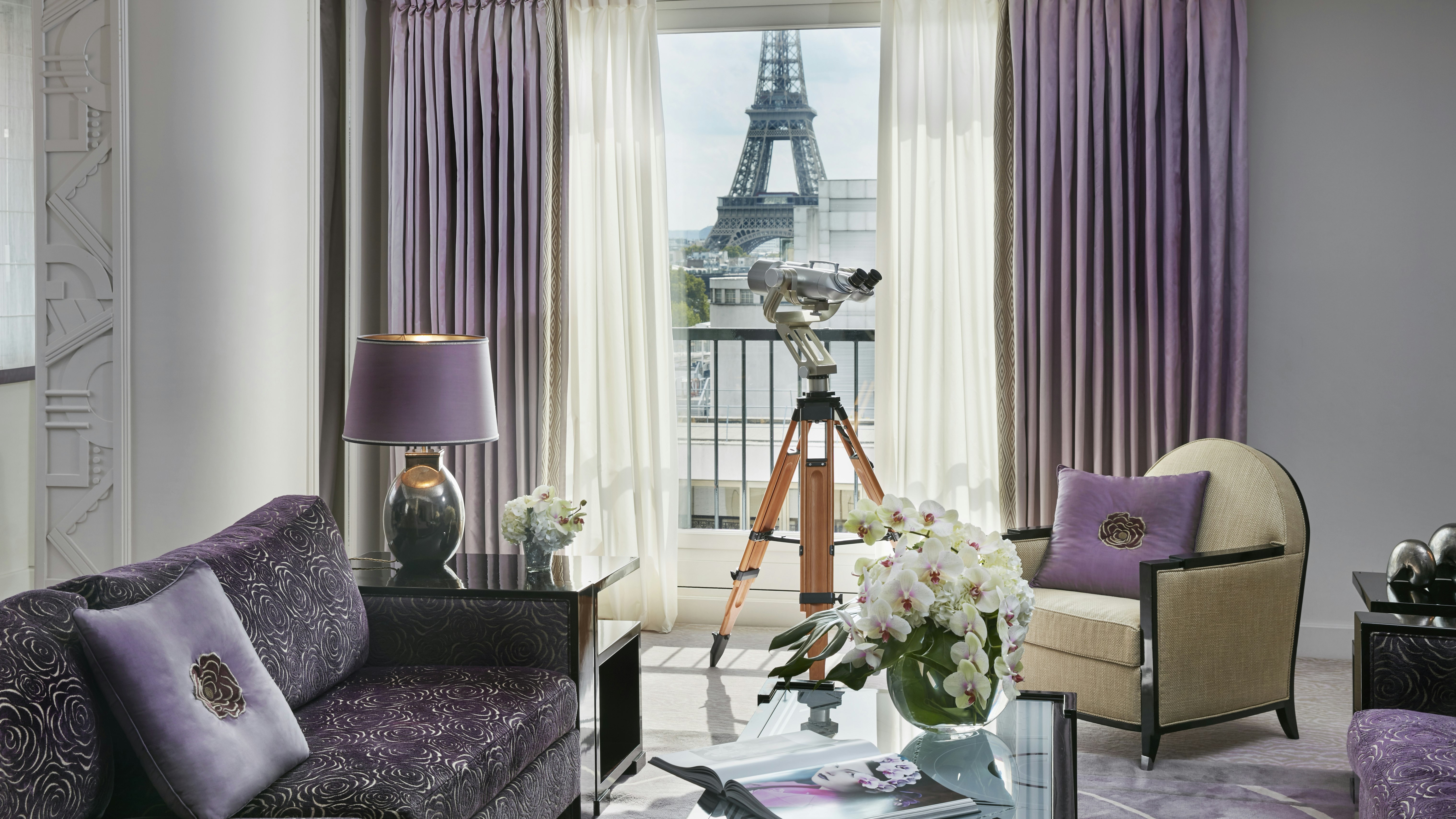 The Eiffel Suite at Hotel Plaza Athenee in Paris is decorated with a luxe purple sofa, lavender curtains, a grey egg-shaped lamp with a lavender shade, a cream chair with lavender throw pillow, and a coffee table with big white blooms in a green vase. Through the window is a view of the bottom portion of the Eiffel tower, with a telescope positioned for easier viewing