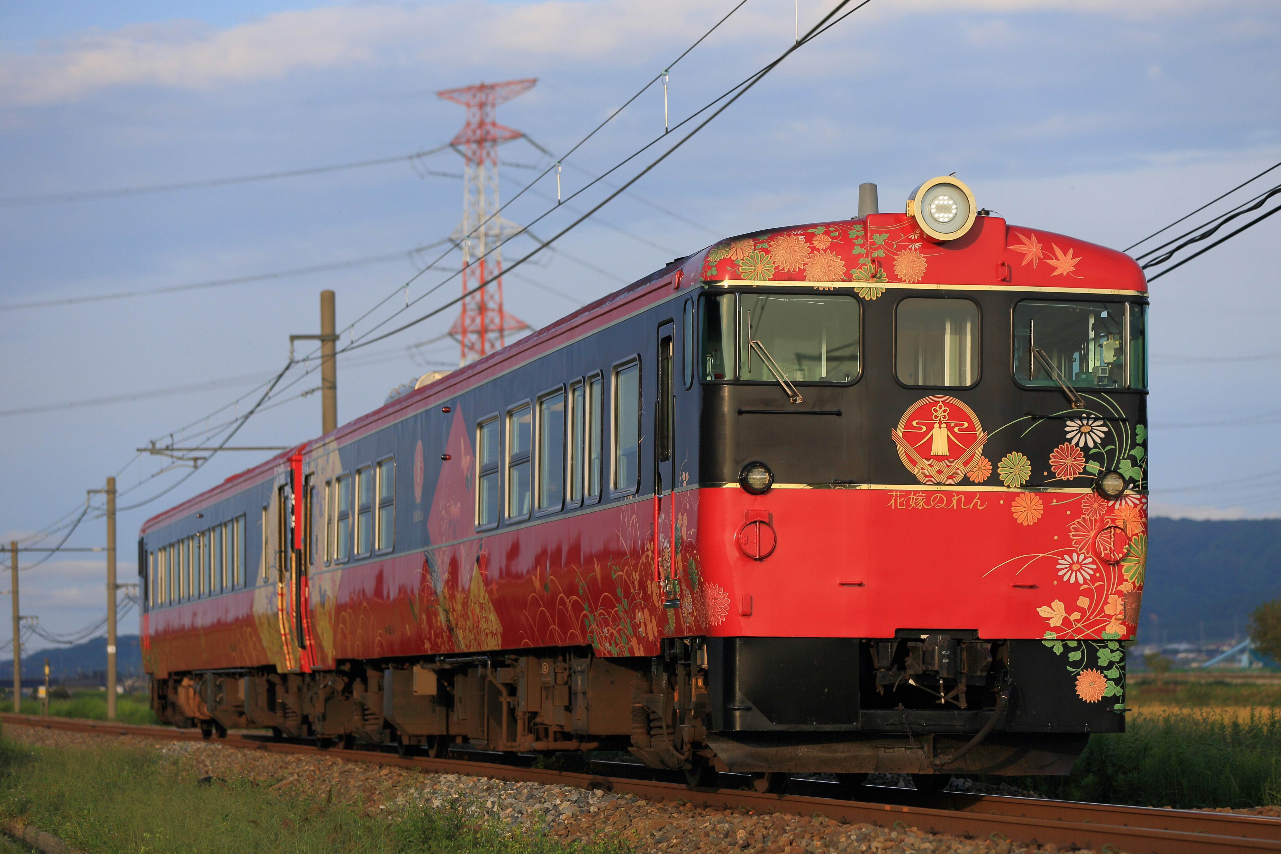 A red train runs along tracks through the Japanese countryside. Green fields are visible beyond it.
