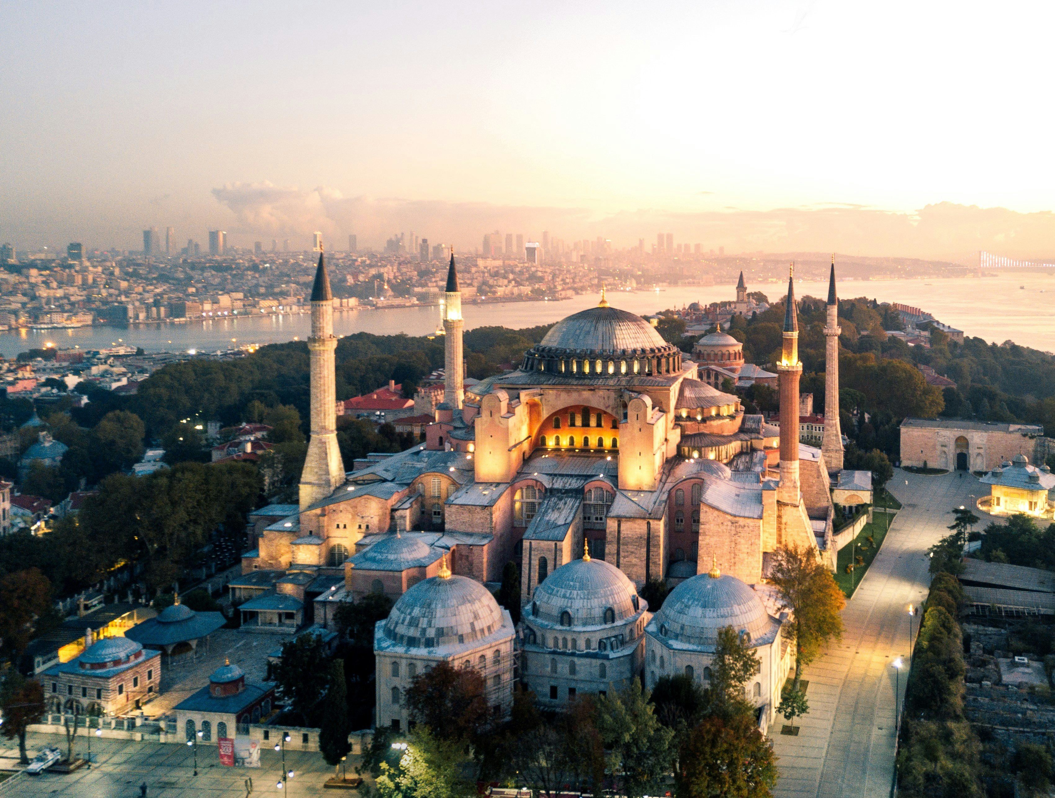 An aerial view of the Hagia Sophia in Istanbul, a former church and mosque and now museum. The building is large and white with four towering minarets on each corner and a domed ceiling. Behind the monument, the Istanbul skyline is visible, including the Bosphorus River that runs through its centre.