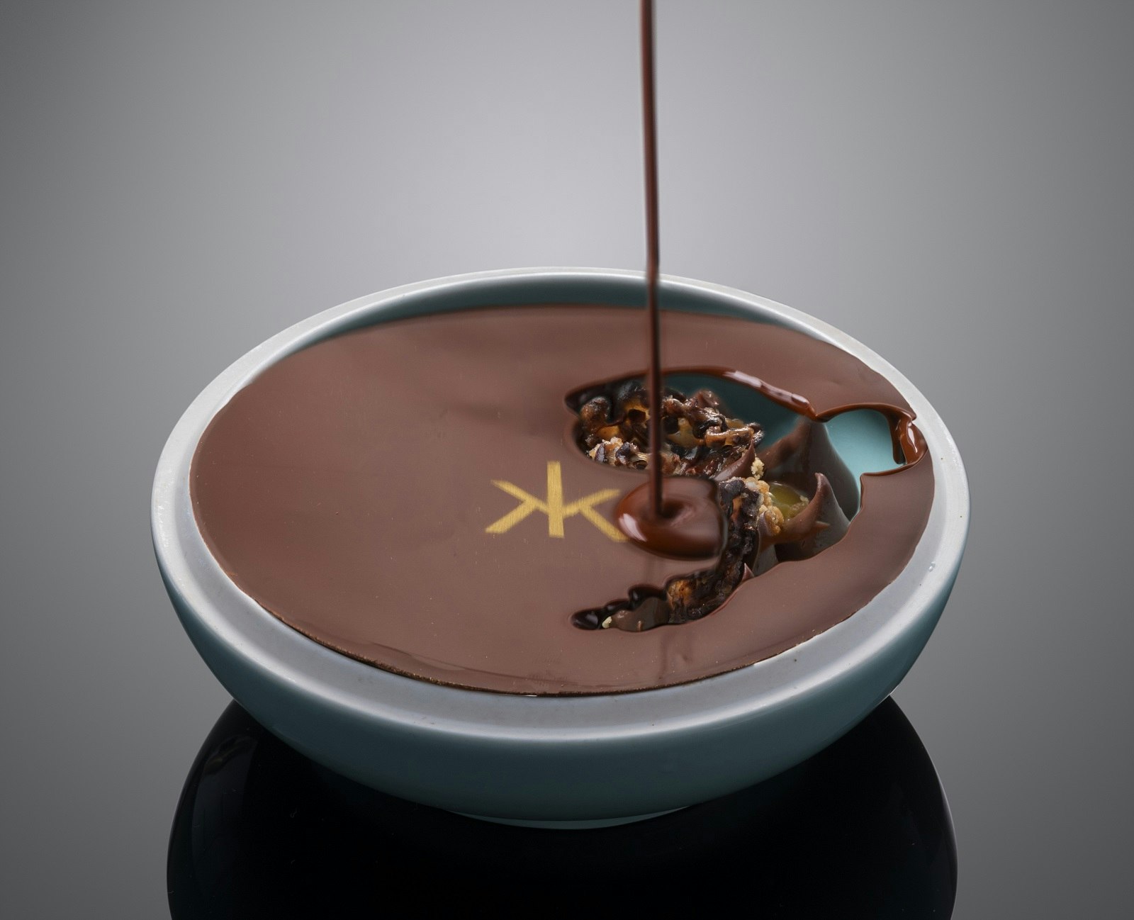 An indulgent Slovenian chocolate pot, featuring the Hakkasan logo in gold. The hard chocolate surface is being drizzled with a hot chocolate sauce to melt the top and reveal the insides. 
