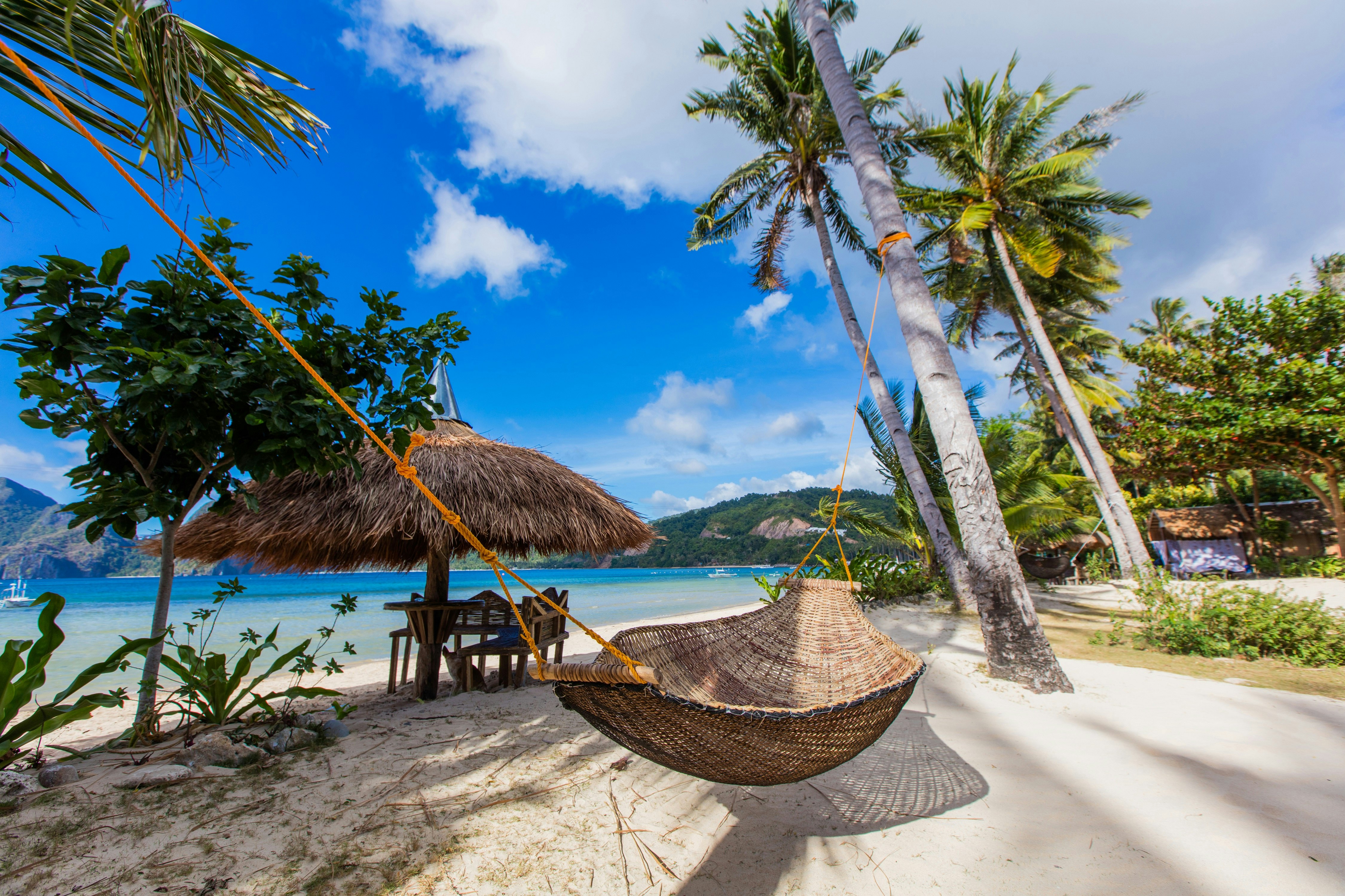A hammock strung between two palm trees next to a beach on Cadlao Island, Palawan, in the Philippines. Behind the hammock a strip of white sand is visible and beyond that a beautiful blue ocean.