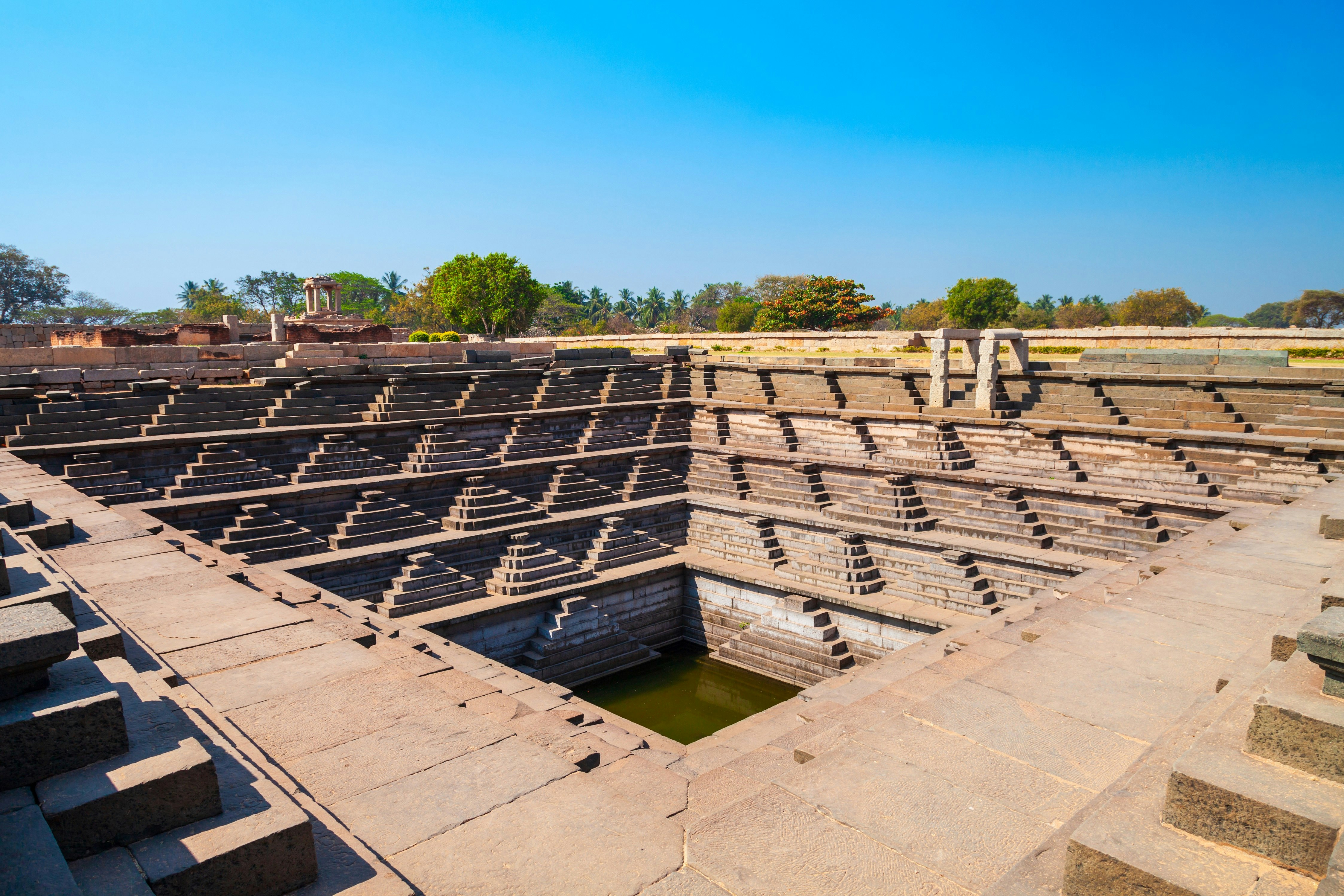 A stepped square water tank at Hampi. Stairs cascade down the walls of the well from all sides, down to a small pool of blue water in the centre.