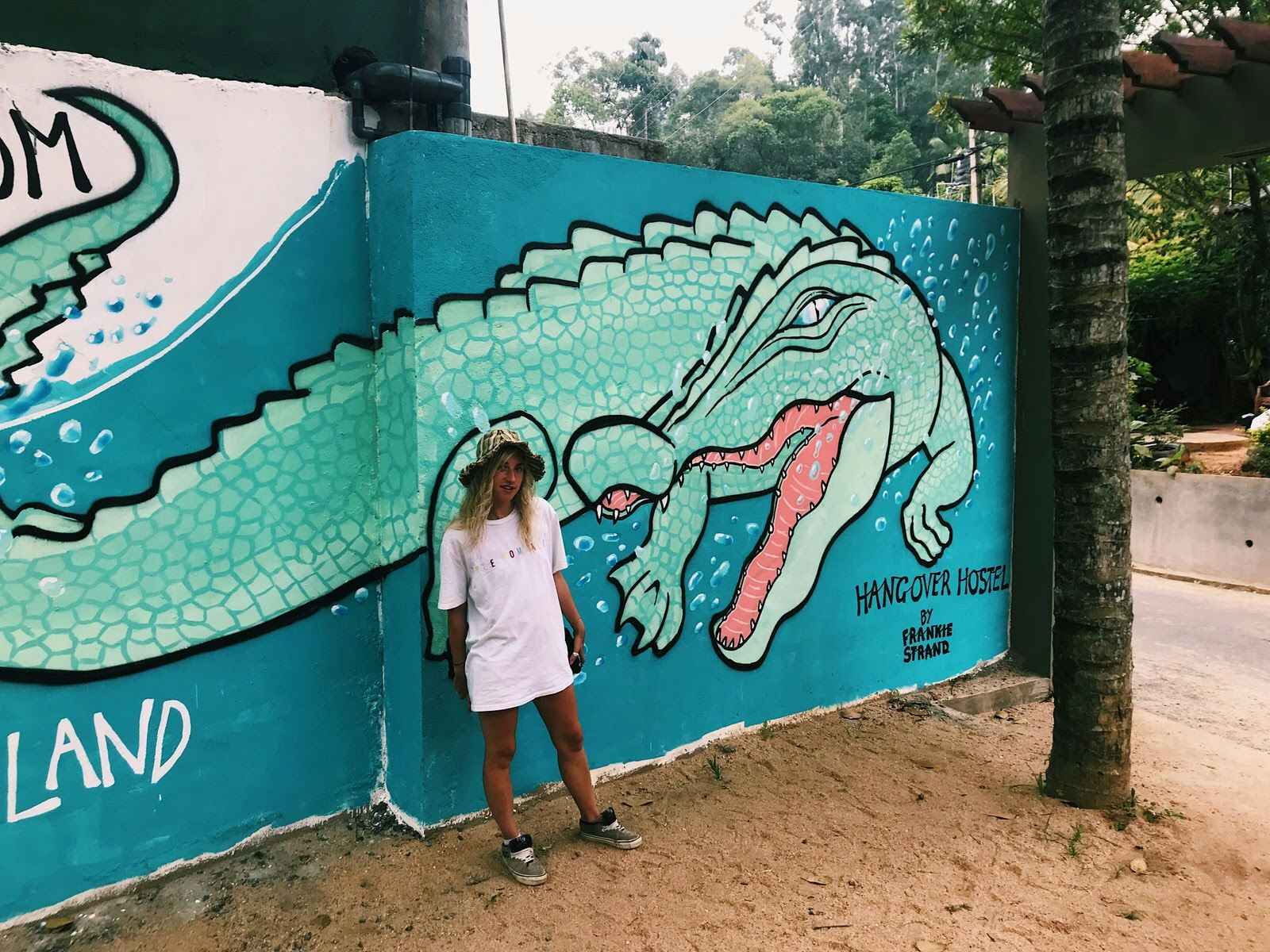 Artist Frankie Strand stands in front of a mural she painted wearing an oversized white t-shirt, trainers and a bucket hat. The mural shows a large alligator turning underwater and has 'HANGOVER HOSTEL' written in the corner.