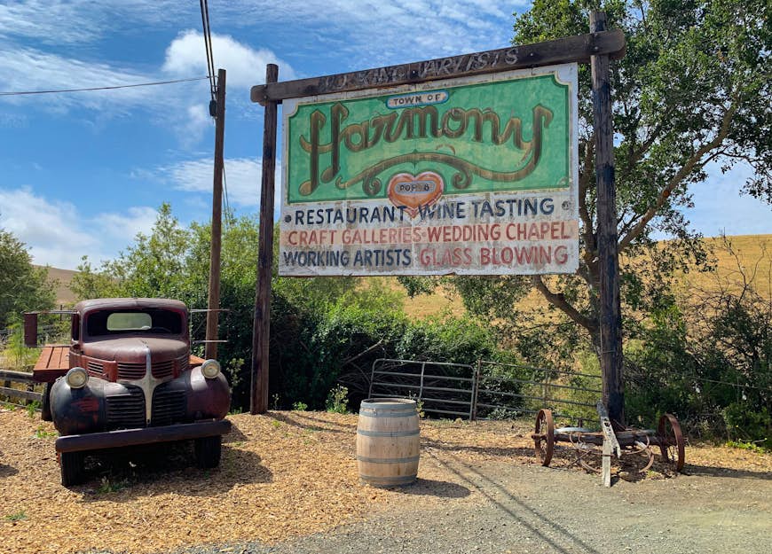 A rusted out old-fashioned truck is parked next to a faded billboard announcing Harmony, California, population 18; California ice cream