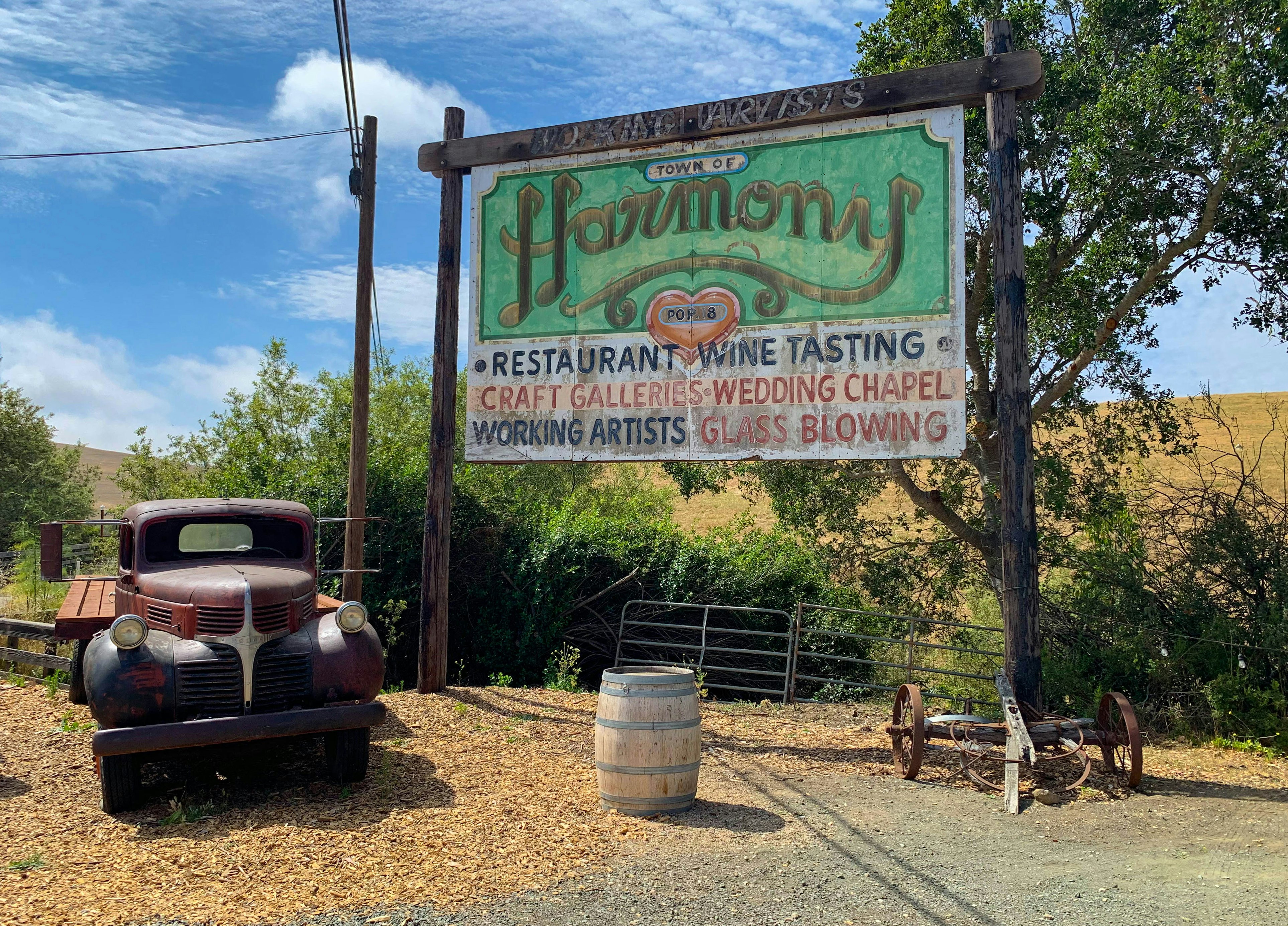 A rusted out old-fashioned truck is parked next to a faded billboard announcing Harmony, California, population 18; California ice cream