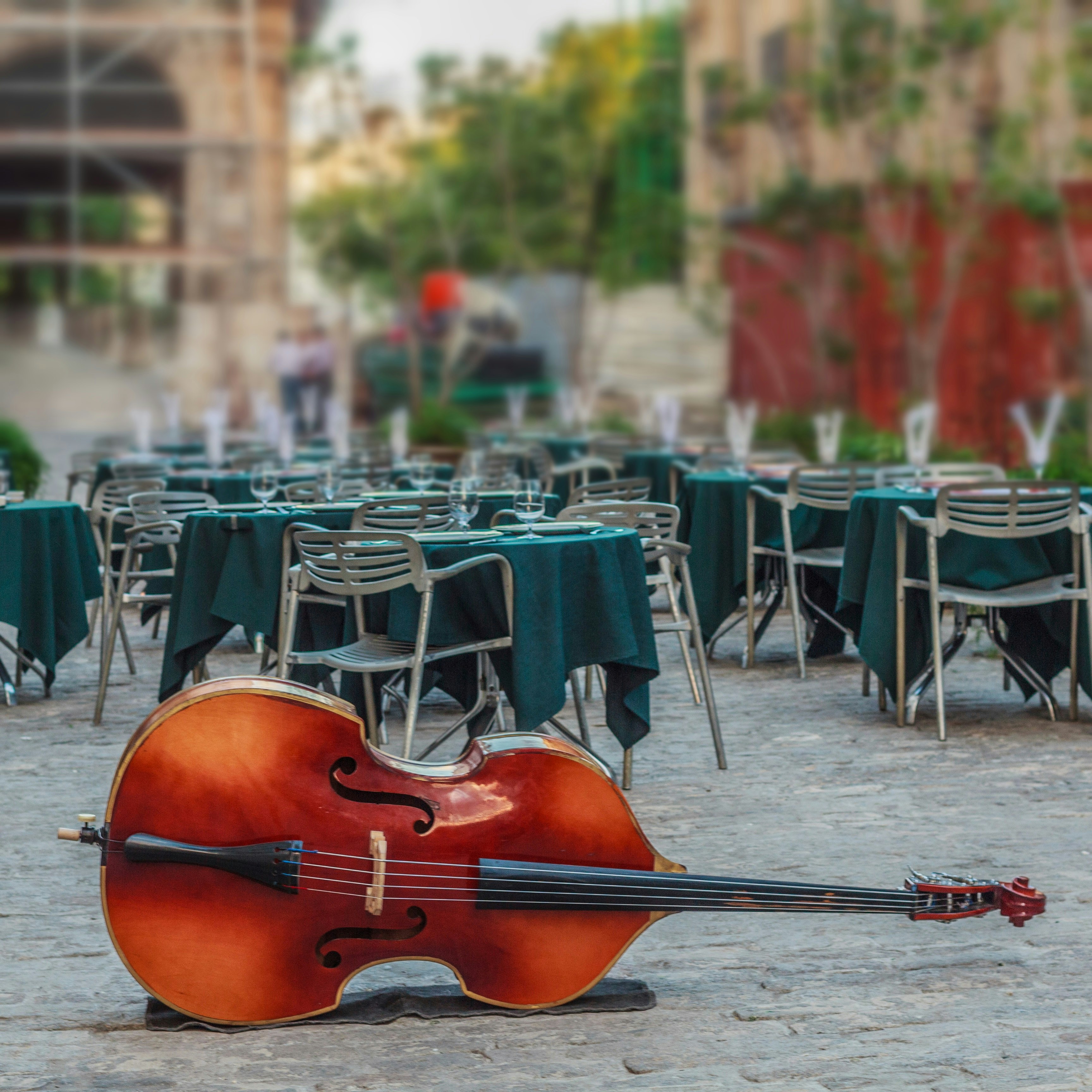 A large cello bass lies on its side in front of a group of tables positioned throughout a open-air cafe; Havana anniversary  