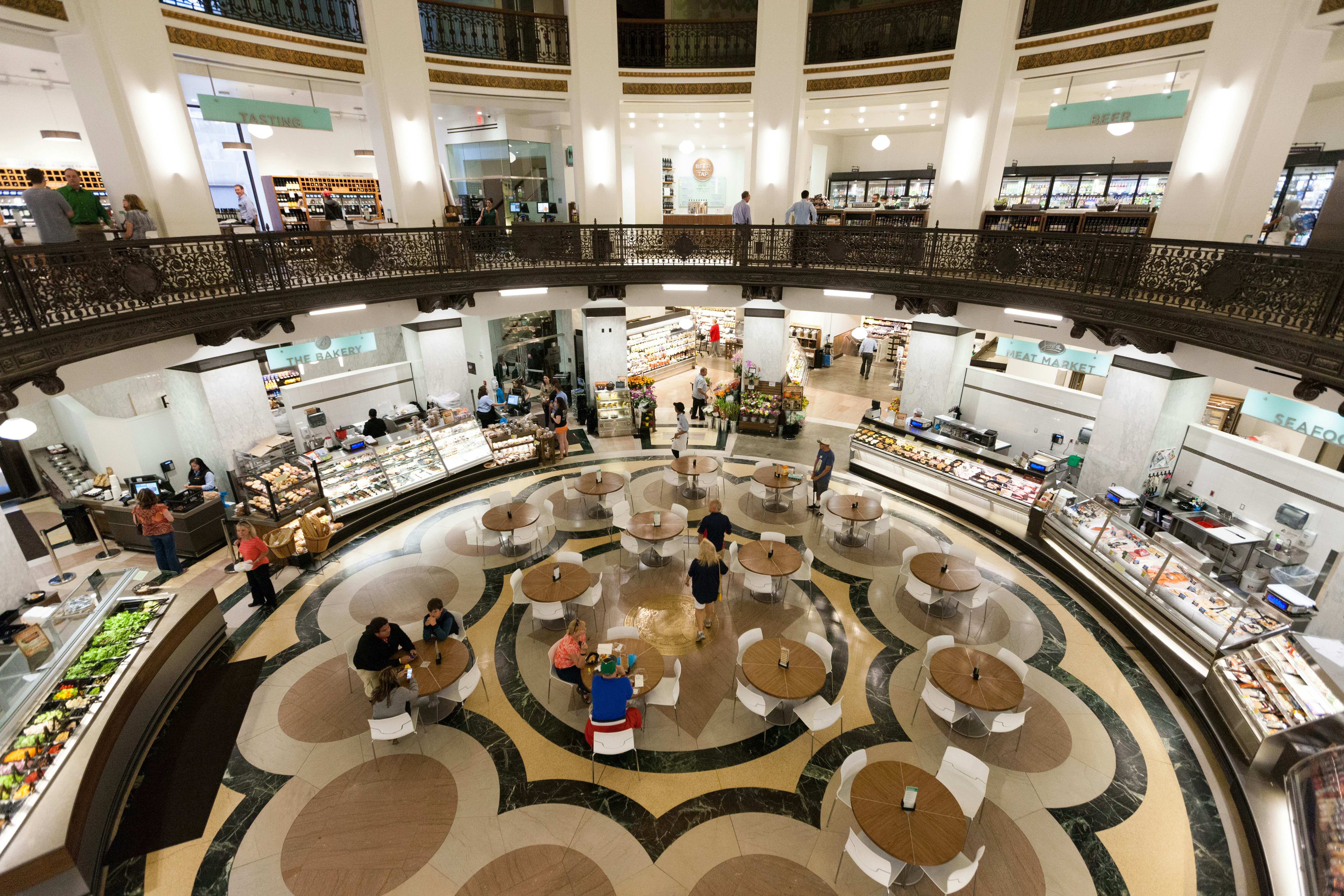 Looking down from a gallery to a cafeteria area inside Heinen's Fine Foods; there are several circular tables and the round space is lined with glass cabinets of food.