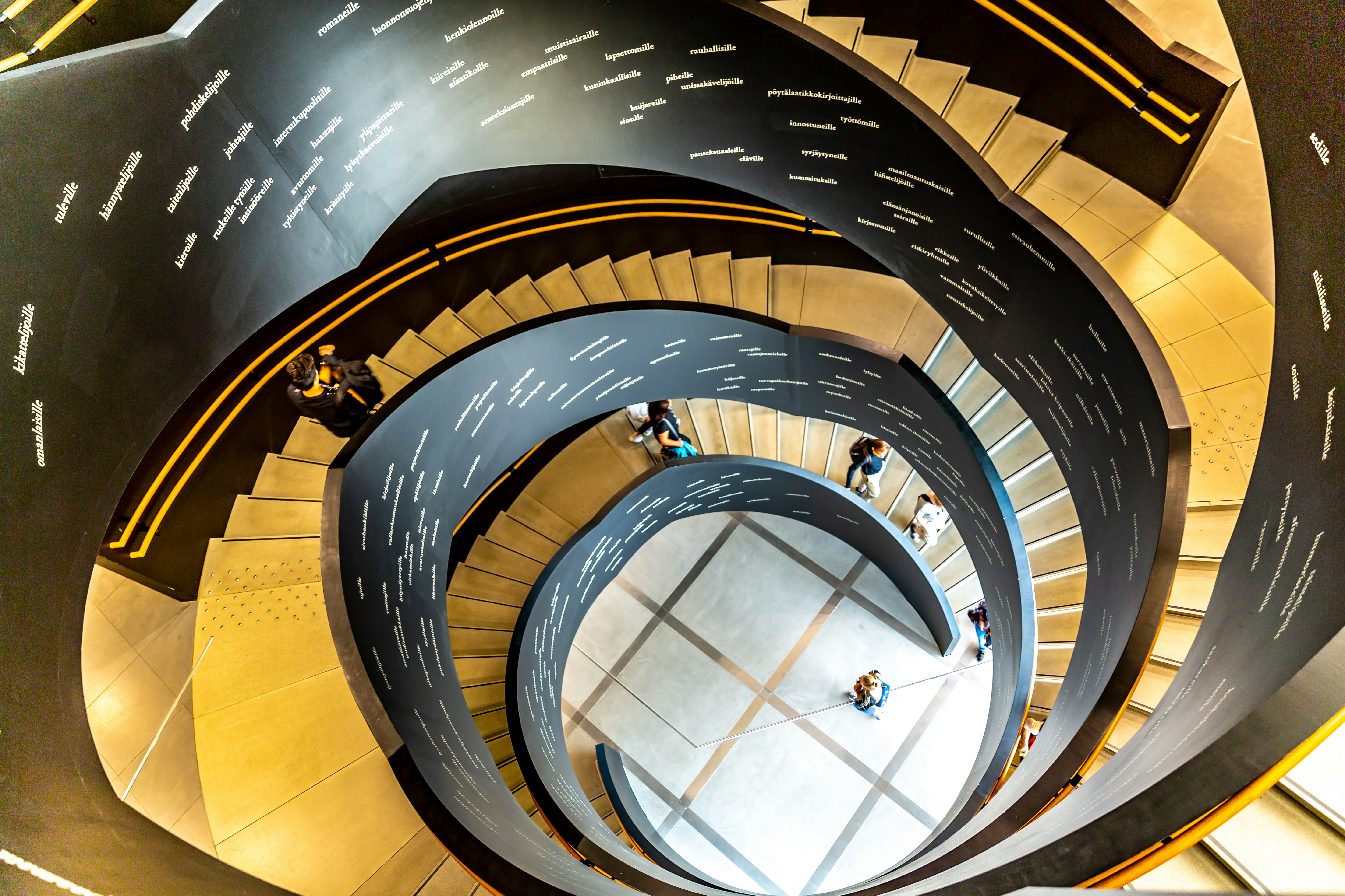 A winding stair in the interior of the New Helsinki city library, which is called Oodi. People are walking up and down the stair, and writing is visible on its inside. 