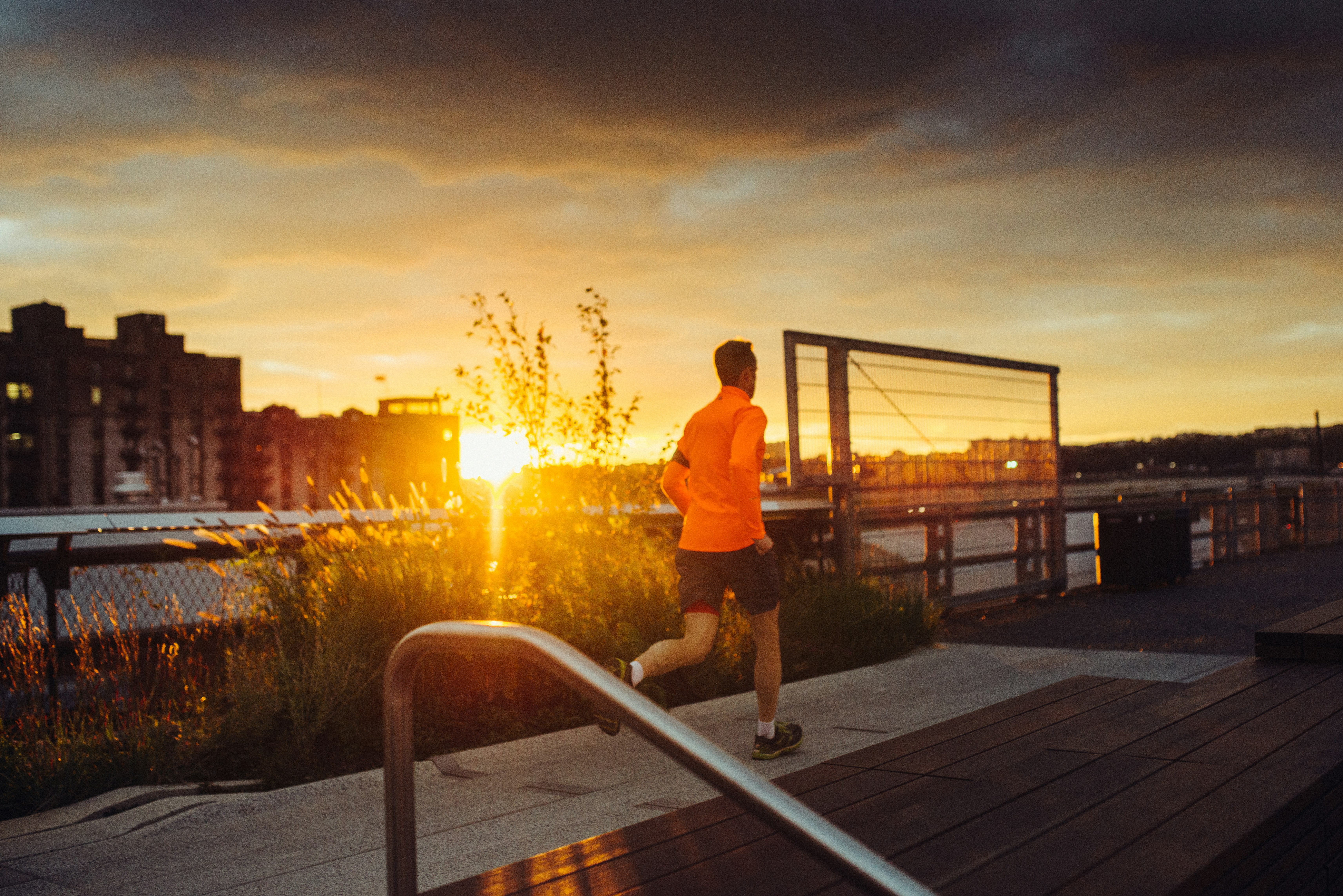 A man in a flourescent orange jacket is running along the New York High Line at sunset.