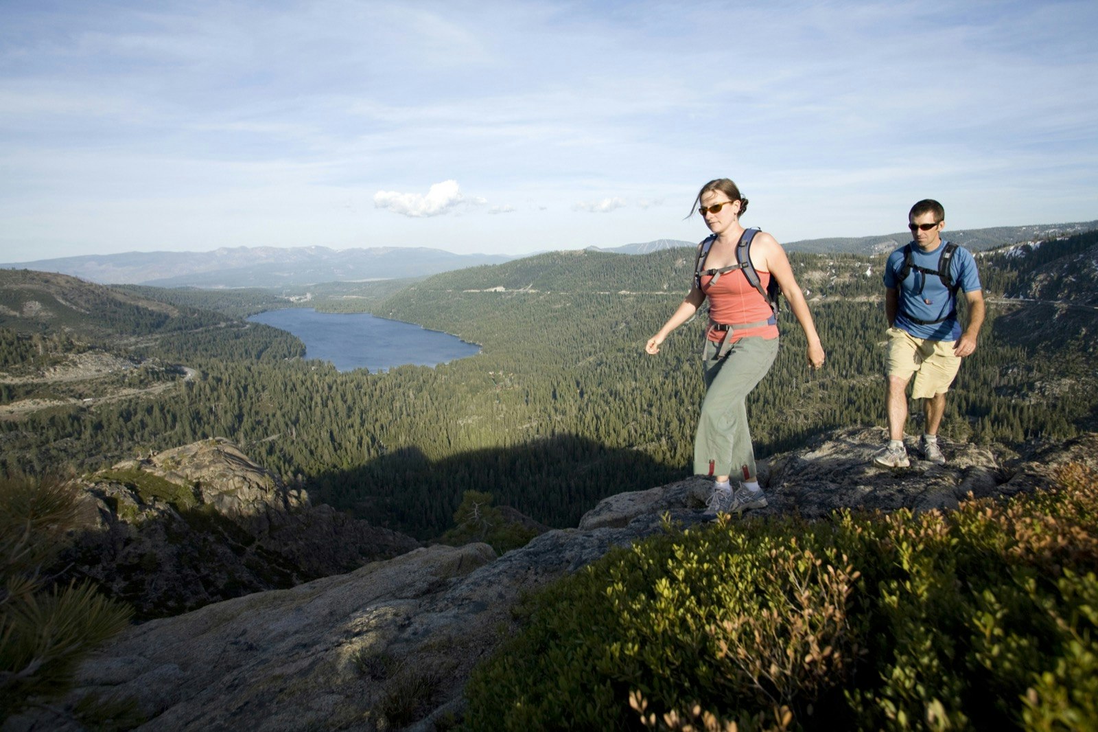 A man and woman hike a ridge line with pine trees and a lake in the distance