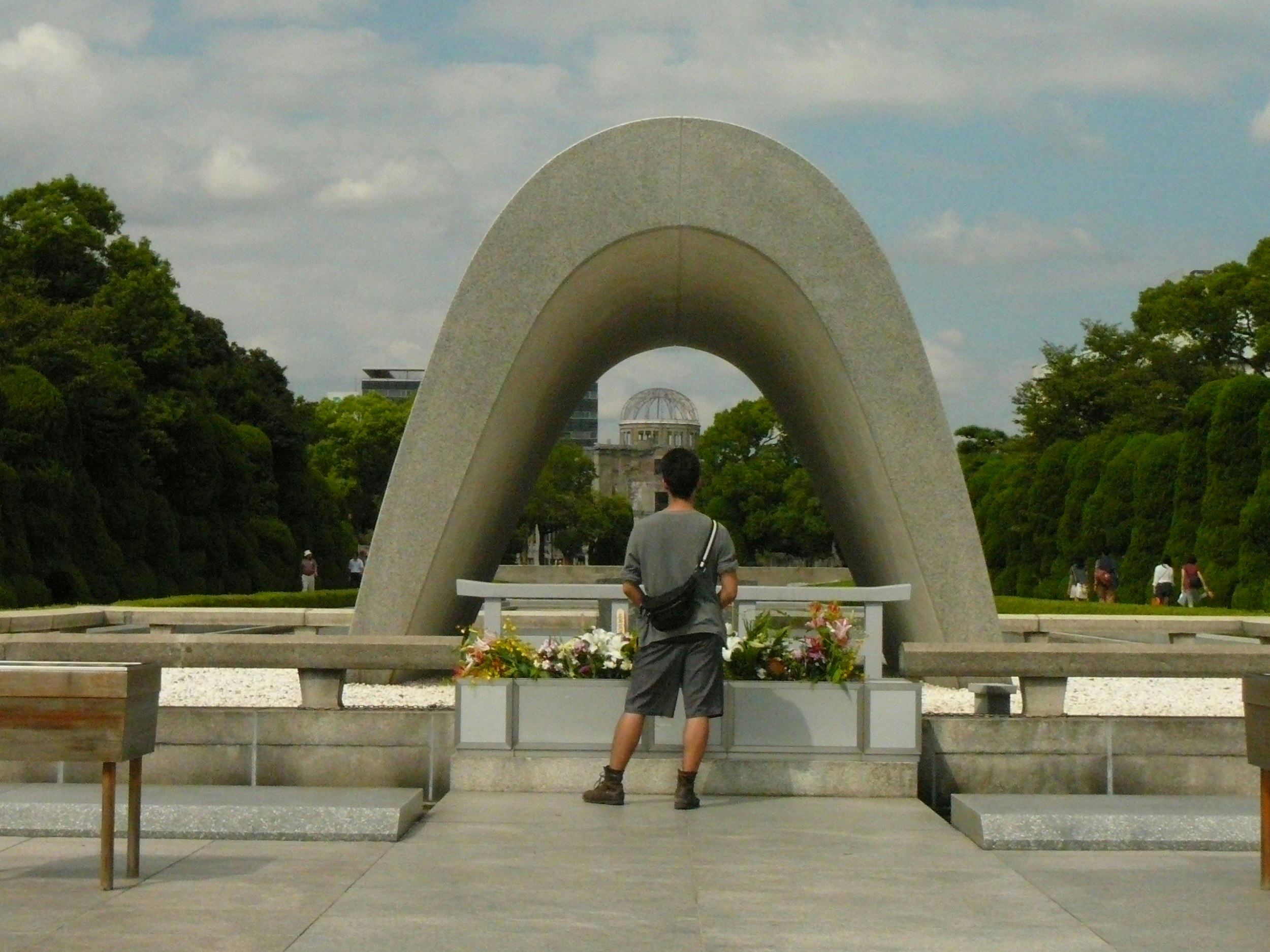 A man stands alone under an archway in a park. In the distance through the arch is what's left of the dome damaged by the Hiroshima nuclear bomb.