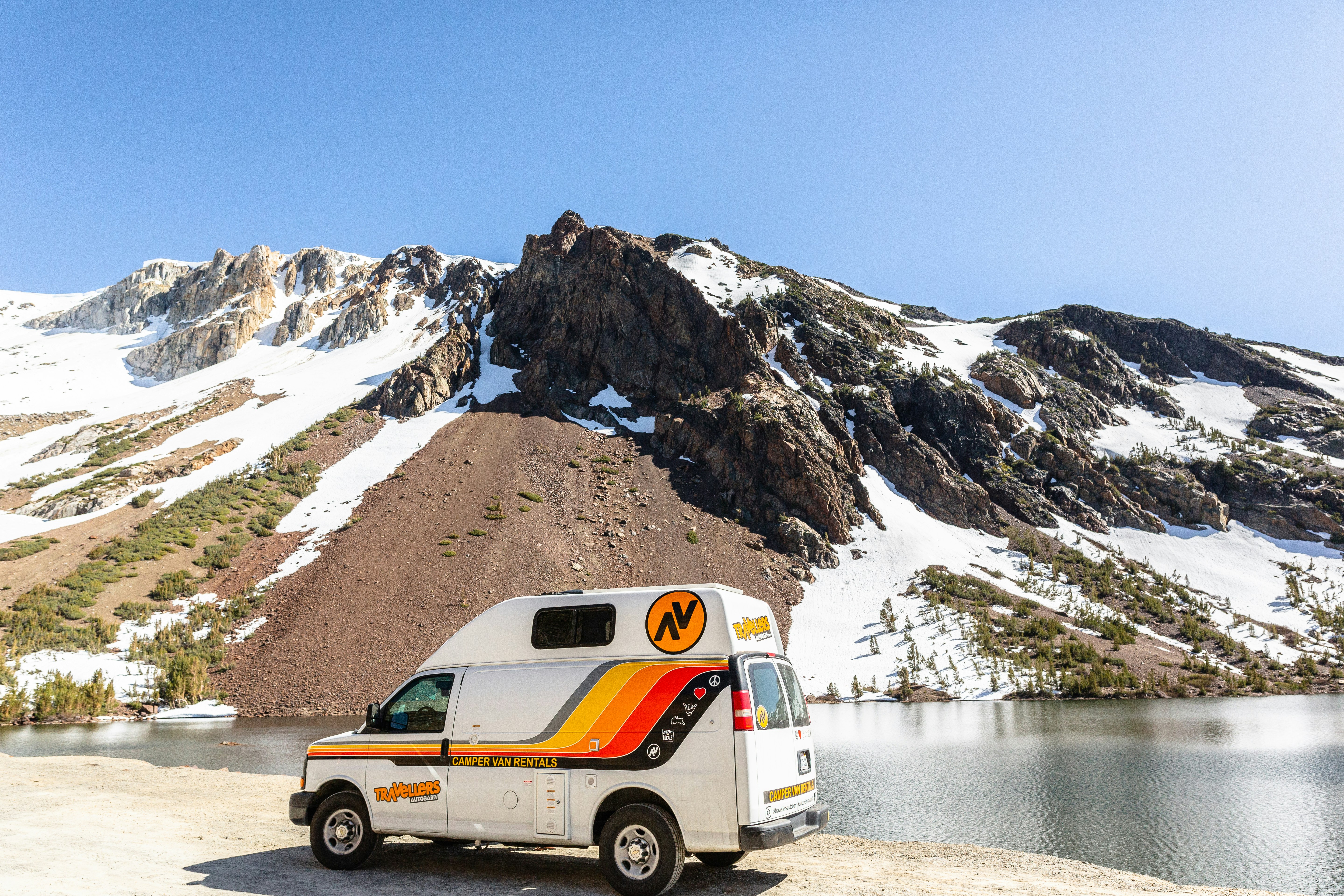 A campervan parked in front of a mountain lake and snow capped rocks