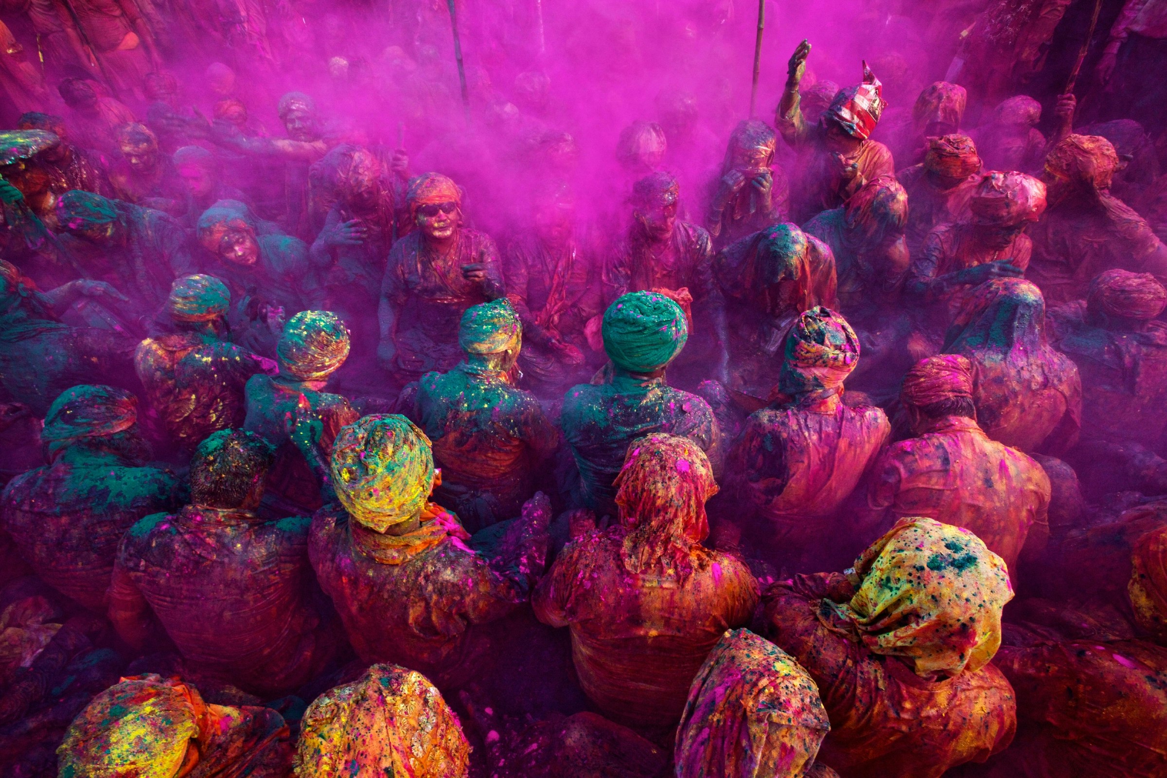 A large group of people, whose clothes are splattered with every colour of the rainbow, sit on the floor cross legged as part of Holi festivities in India.