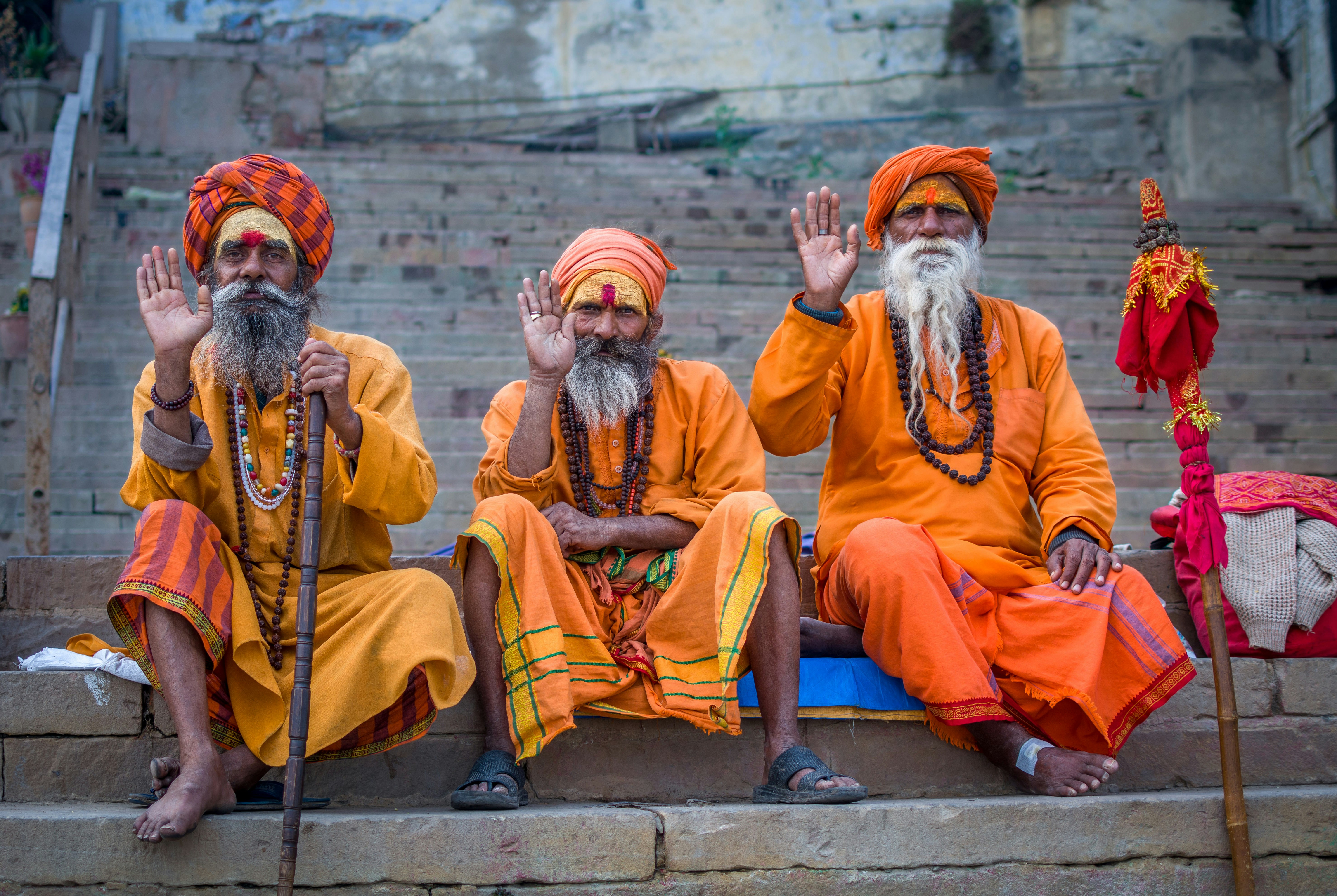 Three holy men dressed in orange robes and turbans, each holding up their right hands, sit on a set on concrete steps in Varanasi, India.
