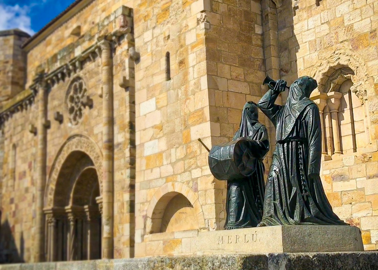 Dark, metallic statues of figures wearing hoods, beating drums and sounding horns, stand in front of the sandstone facade of a Romanesque church in Zamora, Spain