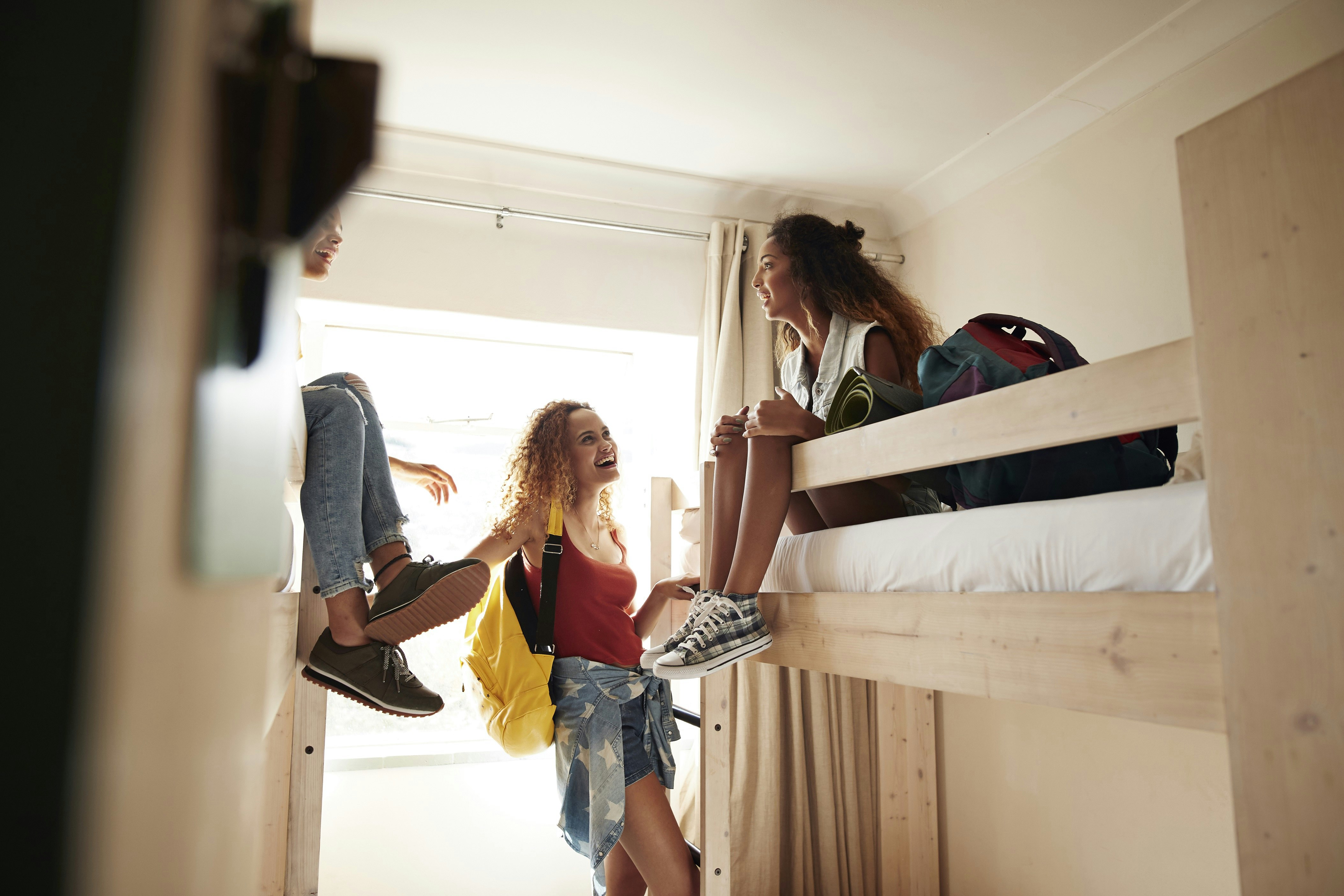 Three young women chat in a hostel dormitory. Two of the girls sit on the top bunk of opposite beds, while the other stands on the floor.