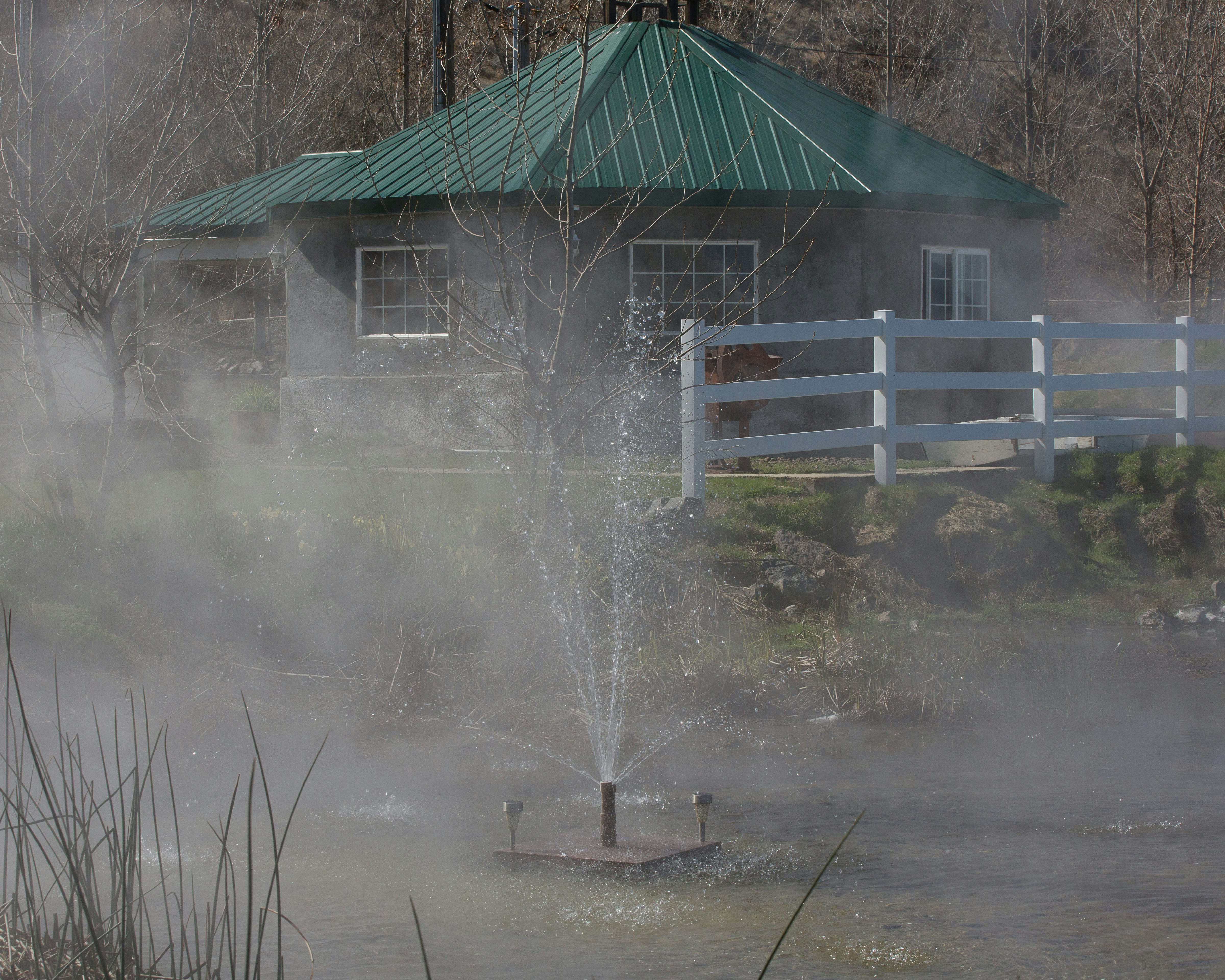 Steam and fountain spray rise from Hot Lake Springs, with a brown building with a green roof in the background