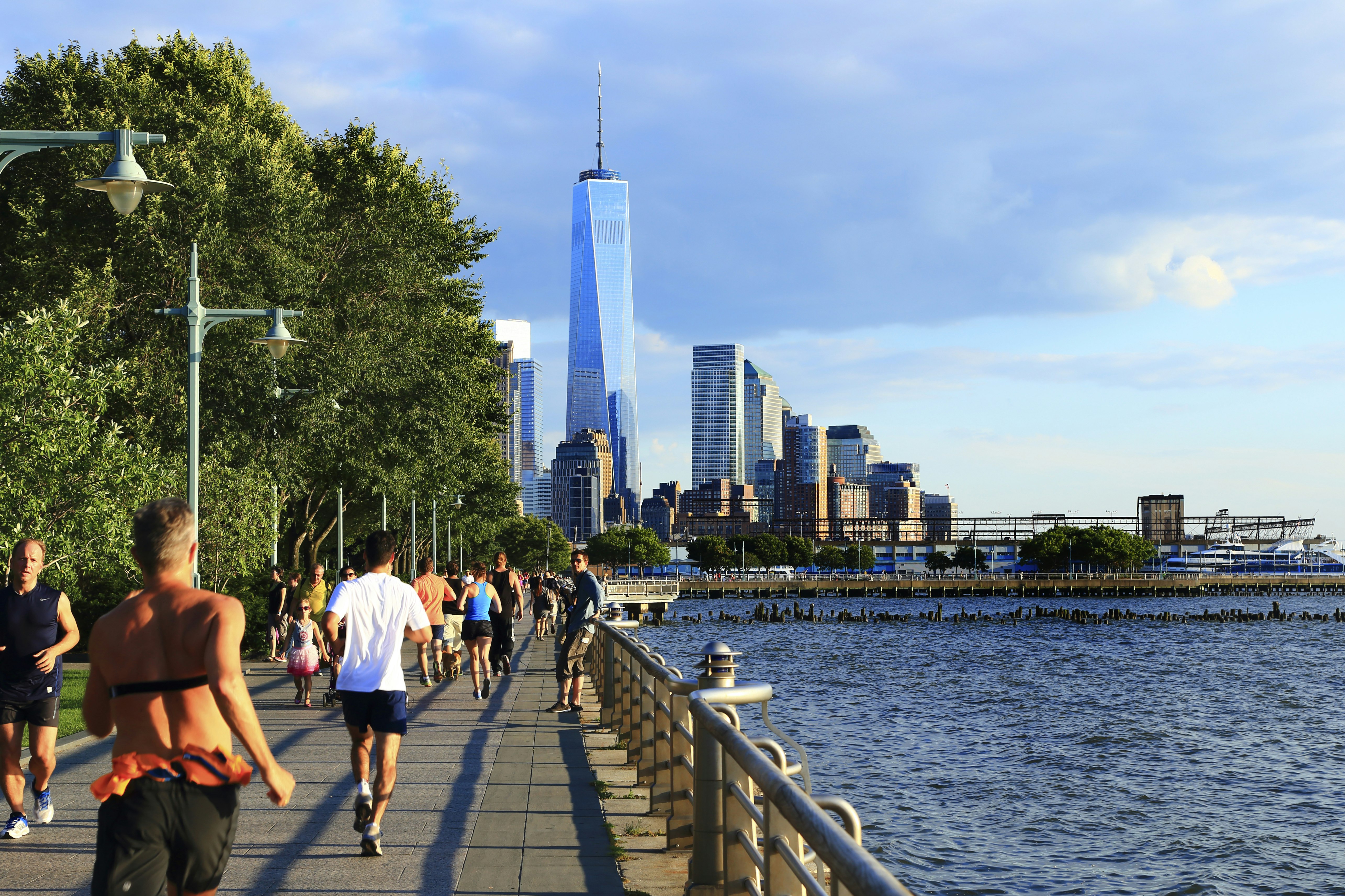 Many people are jogging, running and walking along the Hudson River Greenway. The path runs alongside the Hudson River, and the other side is line with trees. The One World Trade Centre can be seen in the distance.