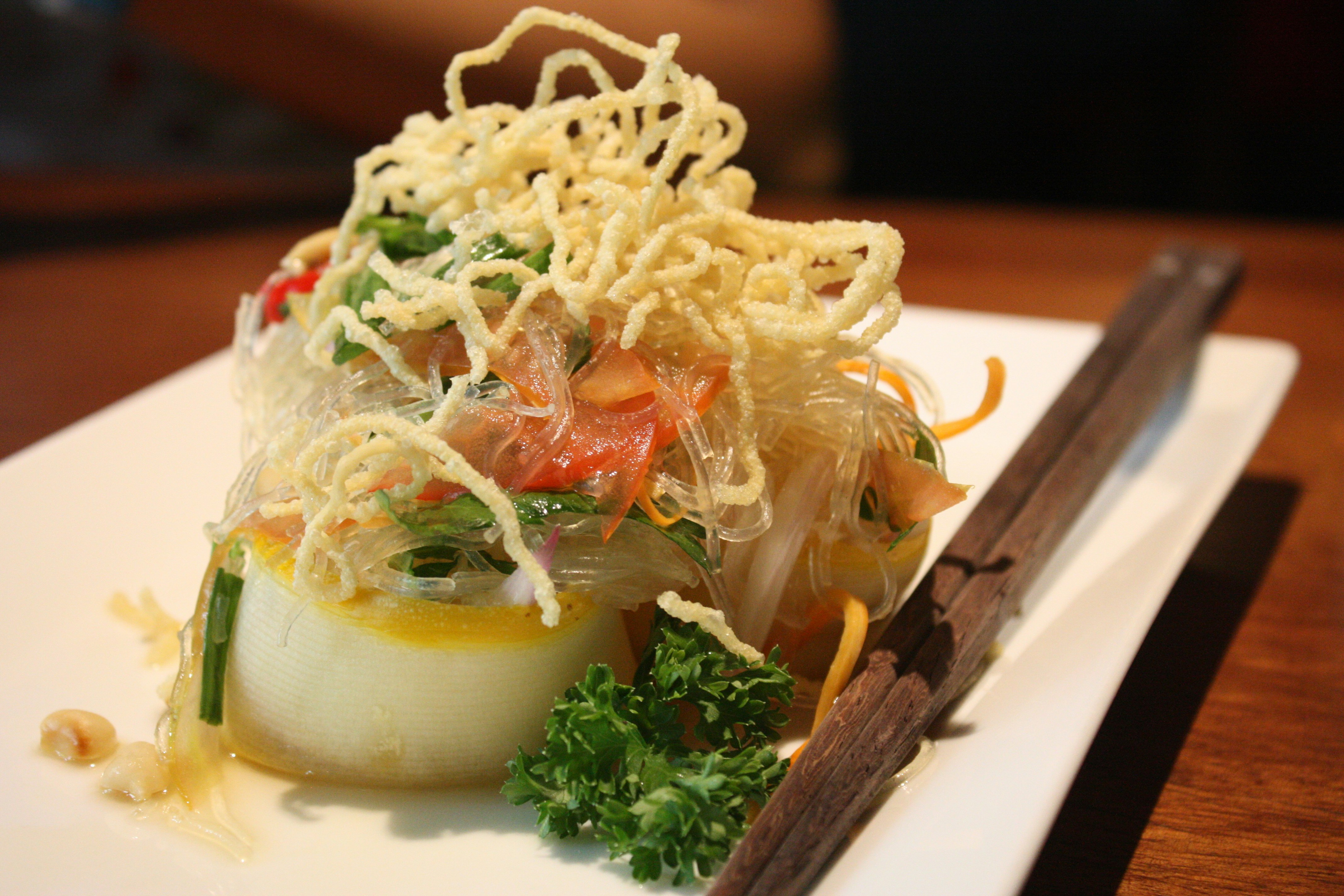 Closeup of medallion-shaped scallop topped with crispy noodles is plated next to wooden chop sticks