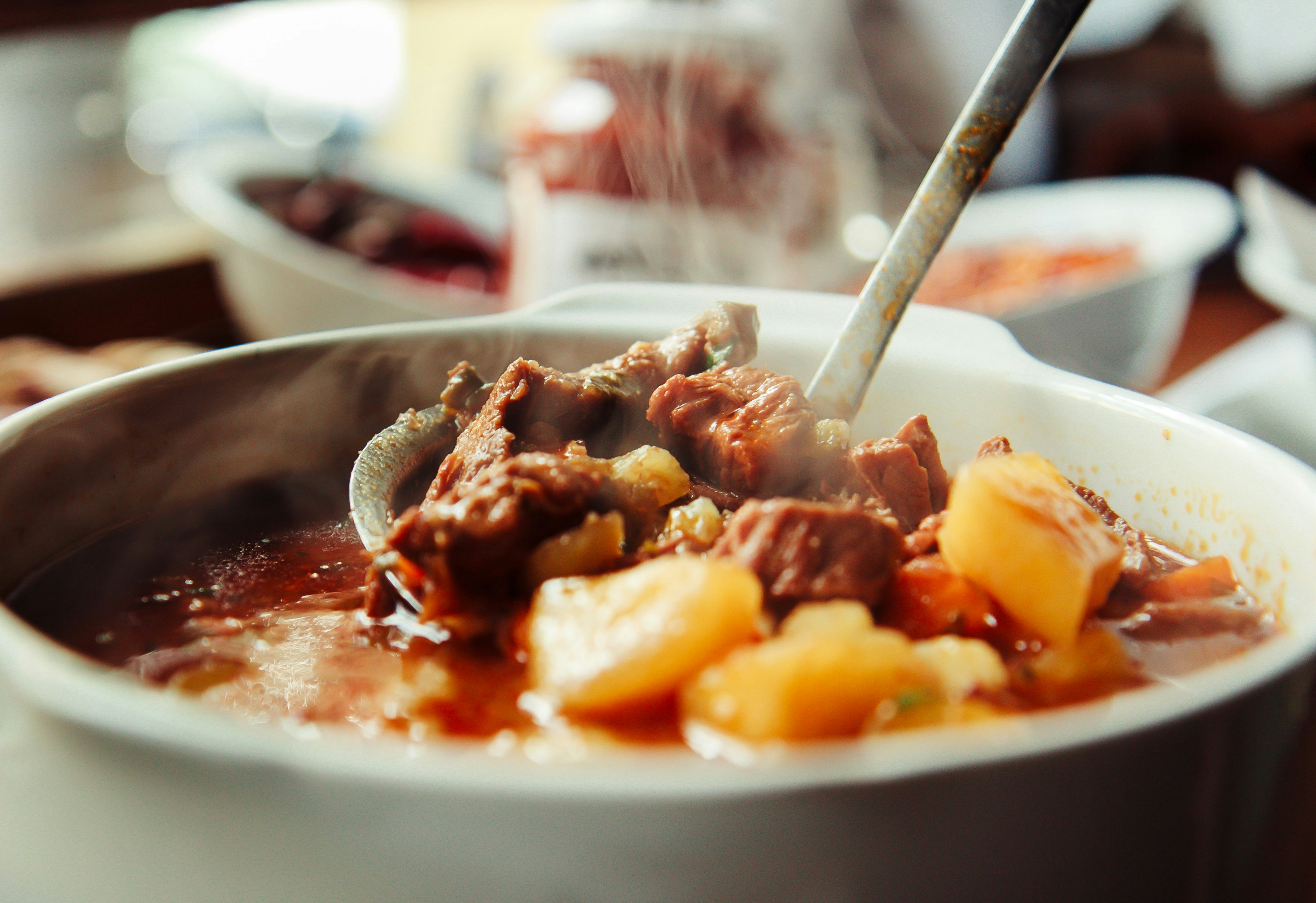 A close-up shot of a metal ladle being dipped into a large bowl of goulash, which appears like a reddish-brown soup with chunks of meat in it.