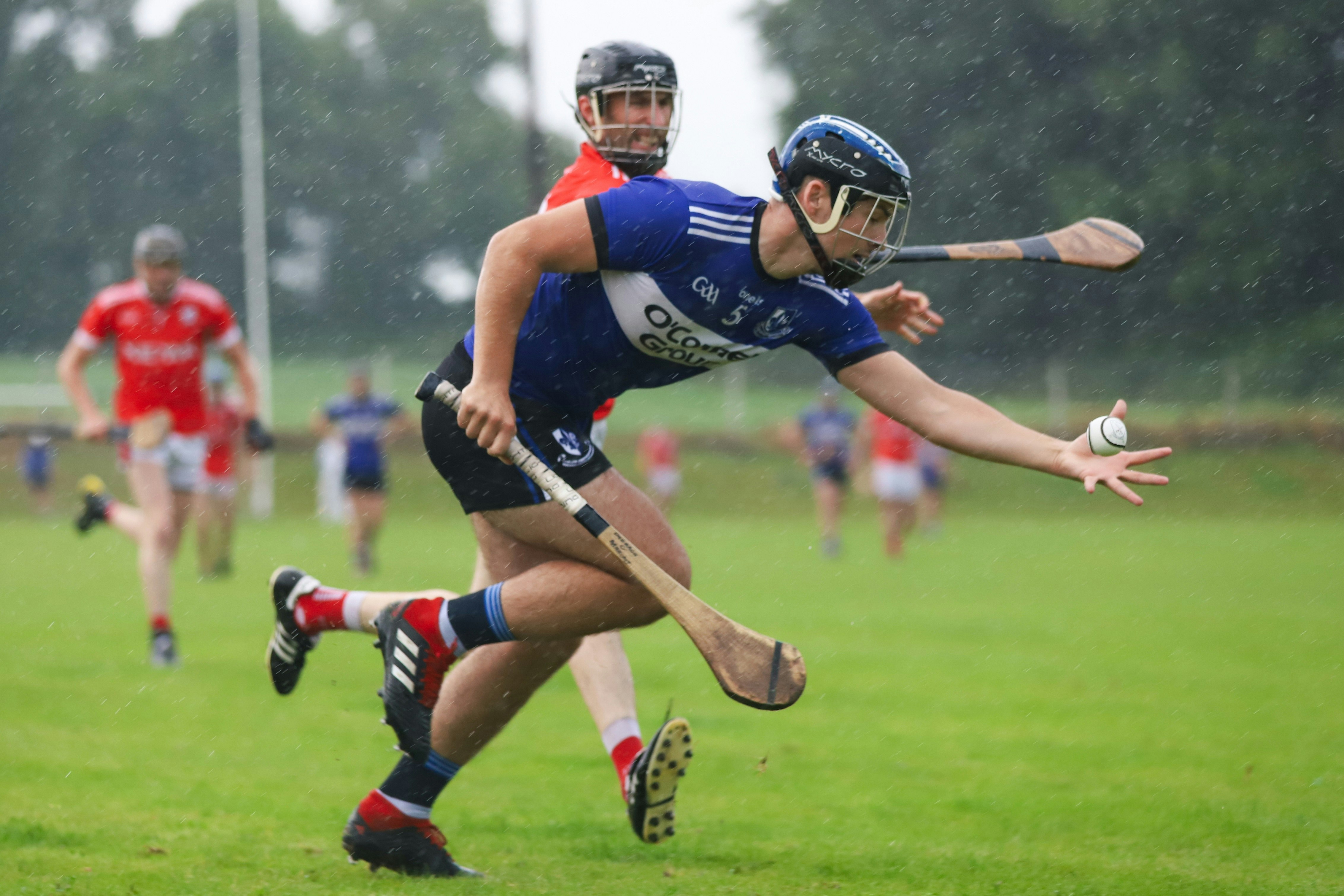 A man in a blue jersey and shorts is holding a hurl and sliotar as a man in a red jersey and shorts attempts to tackle him. 