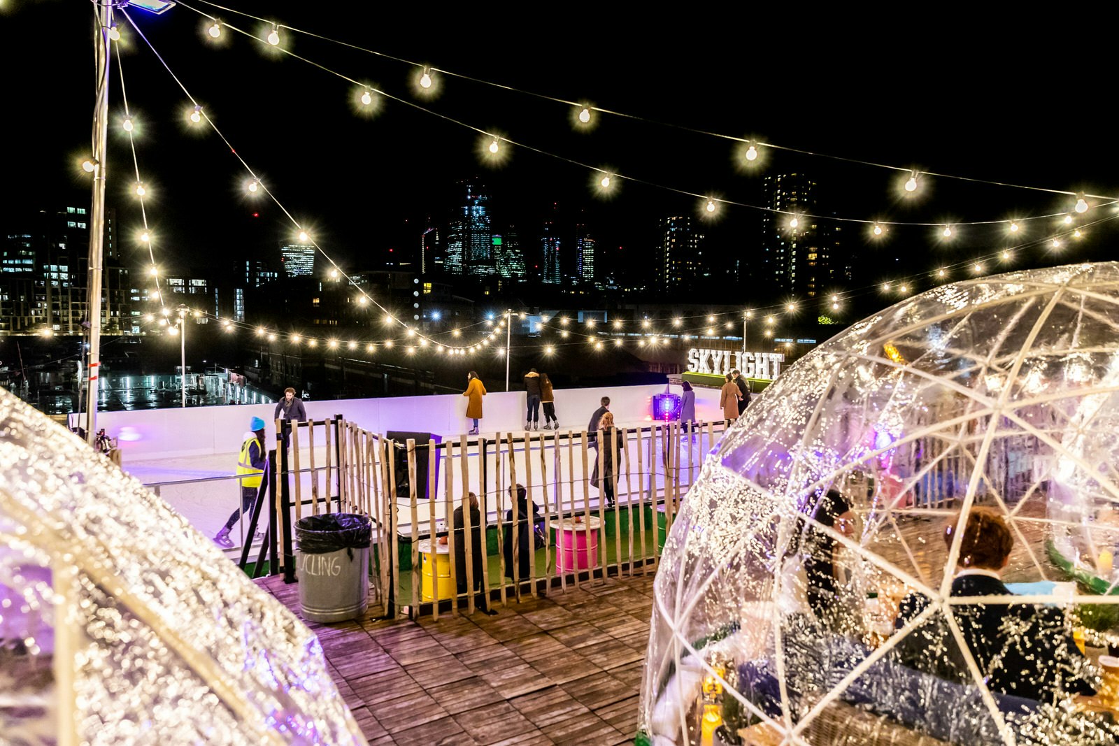 Image taken at night time of the ice rink at Skylight rooftop bar, London. In the distance we can see the lights of many London skyscrapers while a few people skate on the white rink. In the foreground, out of focus, there are plastic igloos full of people drinking and chatting.