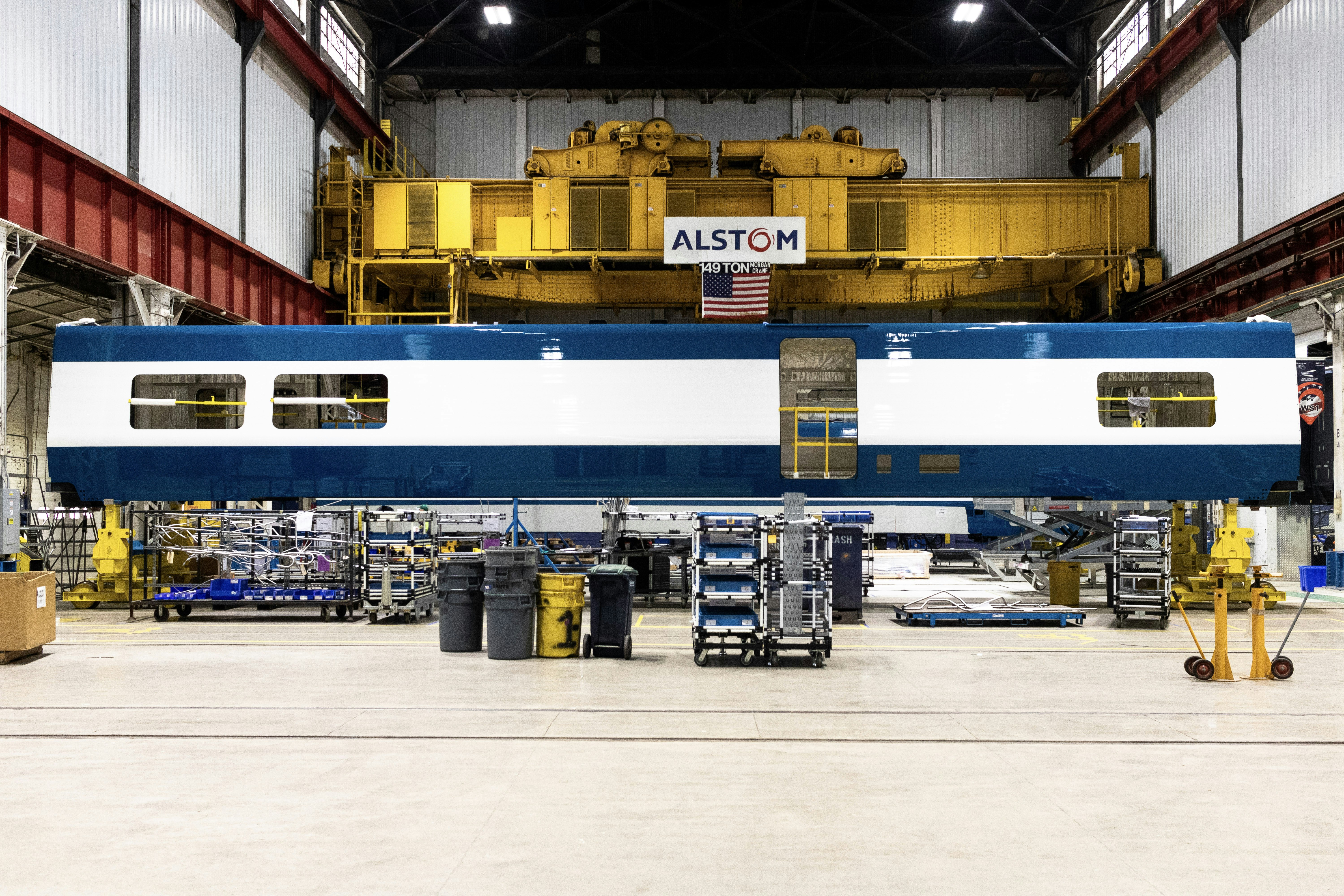 The sleek white and navy striped metal skin of Amtrak's new cafe cars sits on a production line at an Alstom plant, surrounded by bright yellow and red steel industrial equipment, grey and yellow plastic bins, and other manufacturing gear.