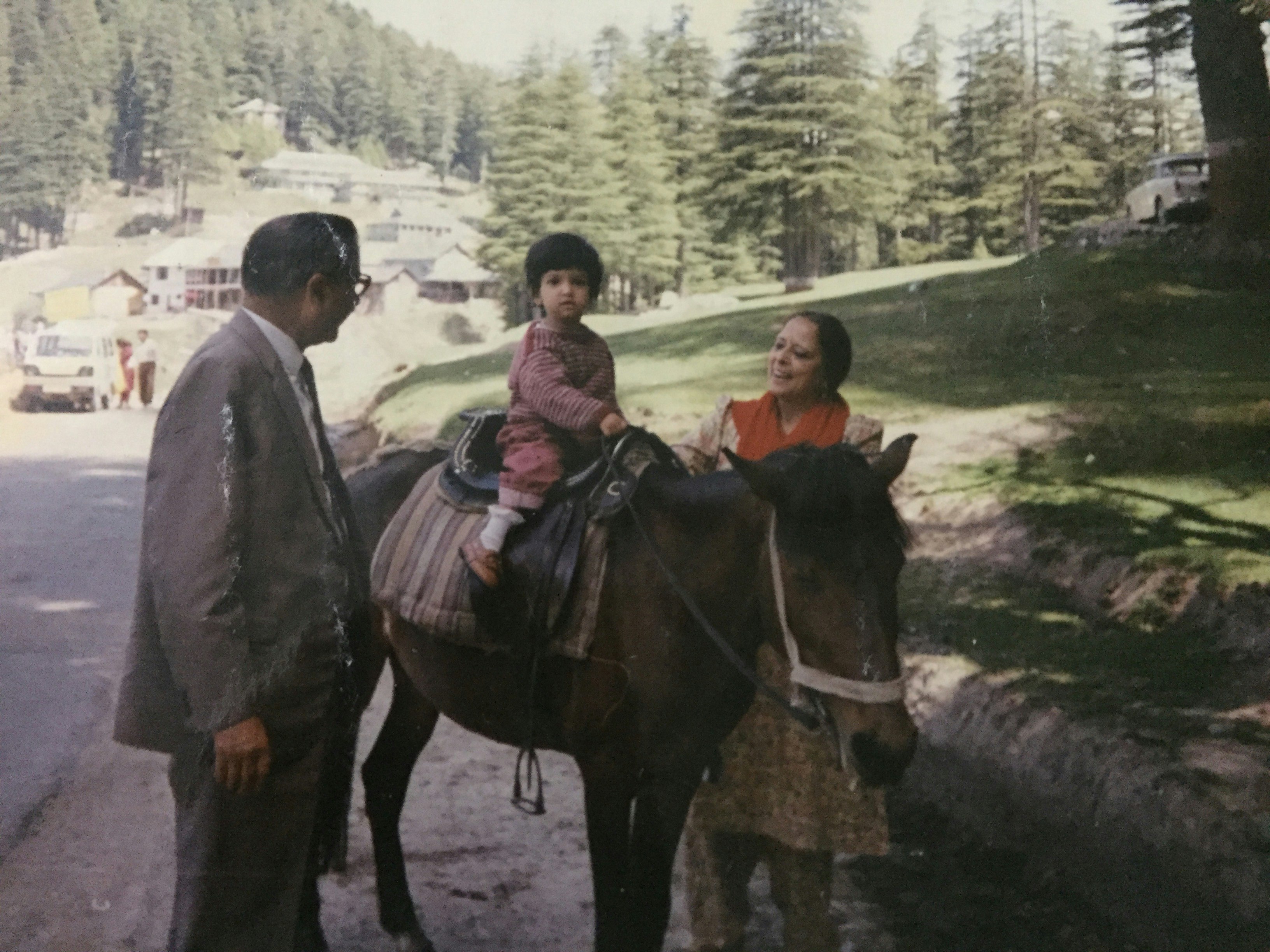 A dated photograph of a young girl riding on the back of a horse, which is being led by two adults. In the background, green forestry and mountain scenery is visible.