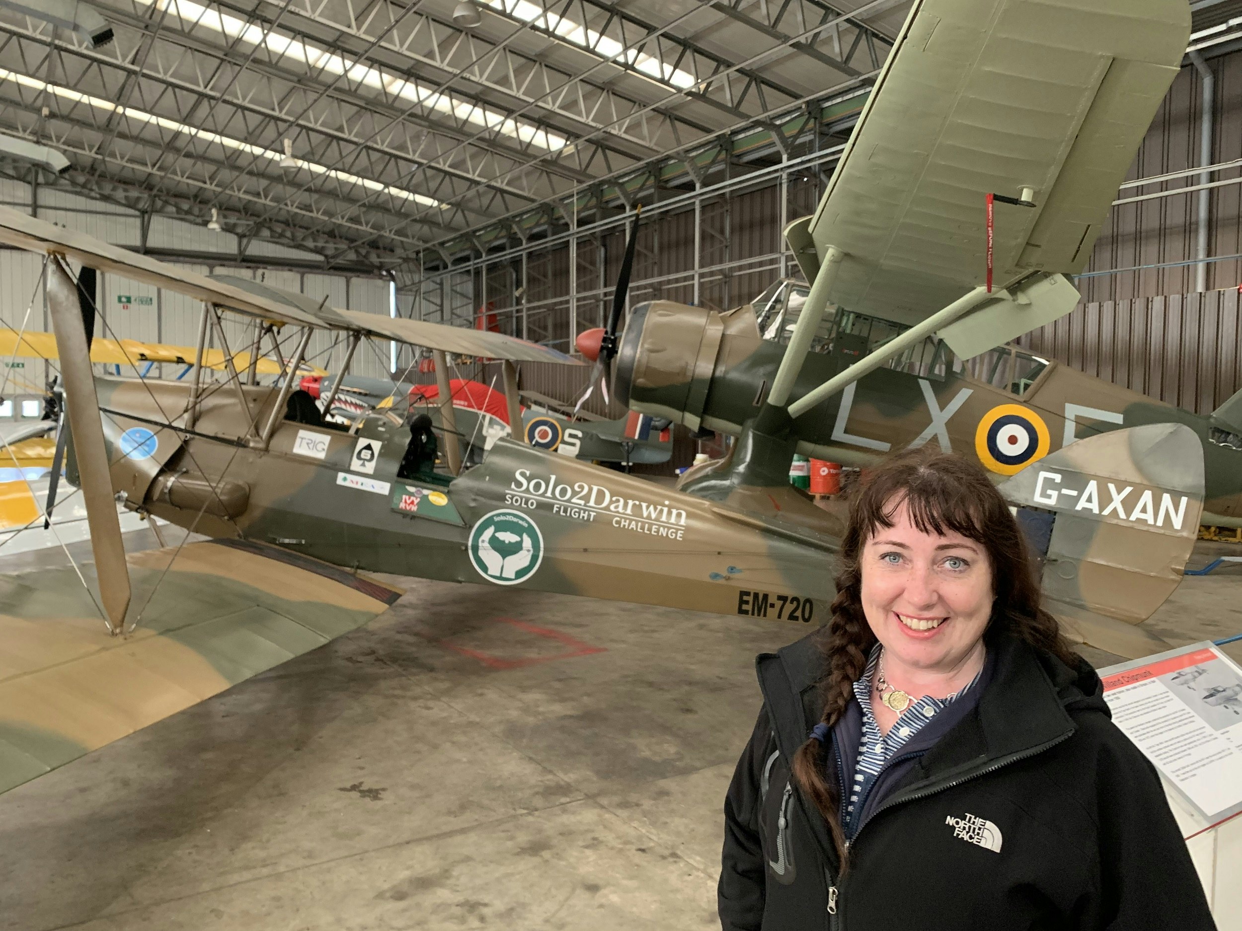 A female pilot stands near her aircraft which is in a hangar. The plane is predominantly brown in colour with several colourful stickers, including one that says Ivy (the name of the plane) and RVH (the initials of the pilot's father) 