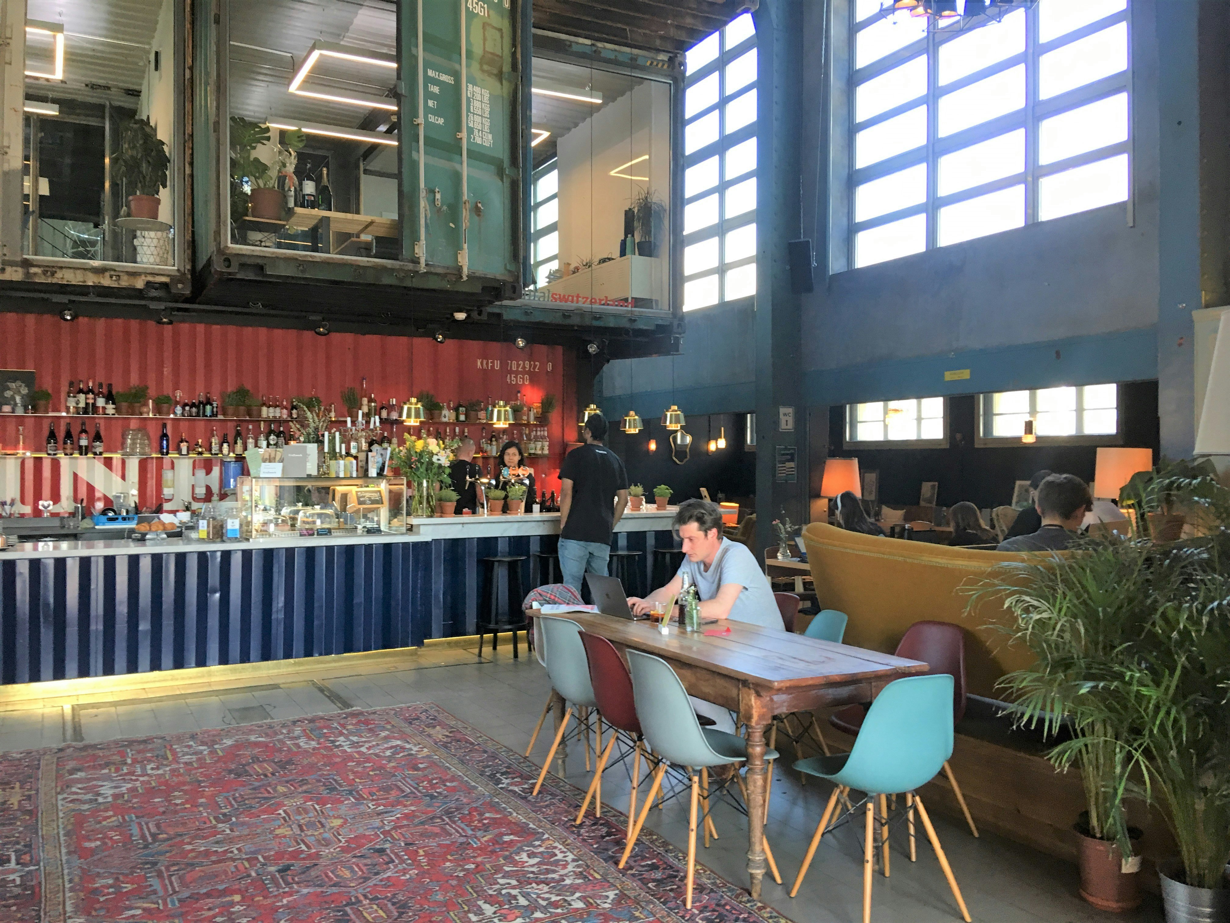 The high-ceilinged interior of Kraftwerk: the bar and some of the walls are made of colourful old shipping containers, while the exterior wall has windows high up; a customer is working at a wooden table next to a large patterned rug covering the floor. 