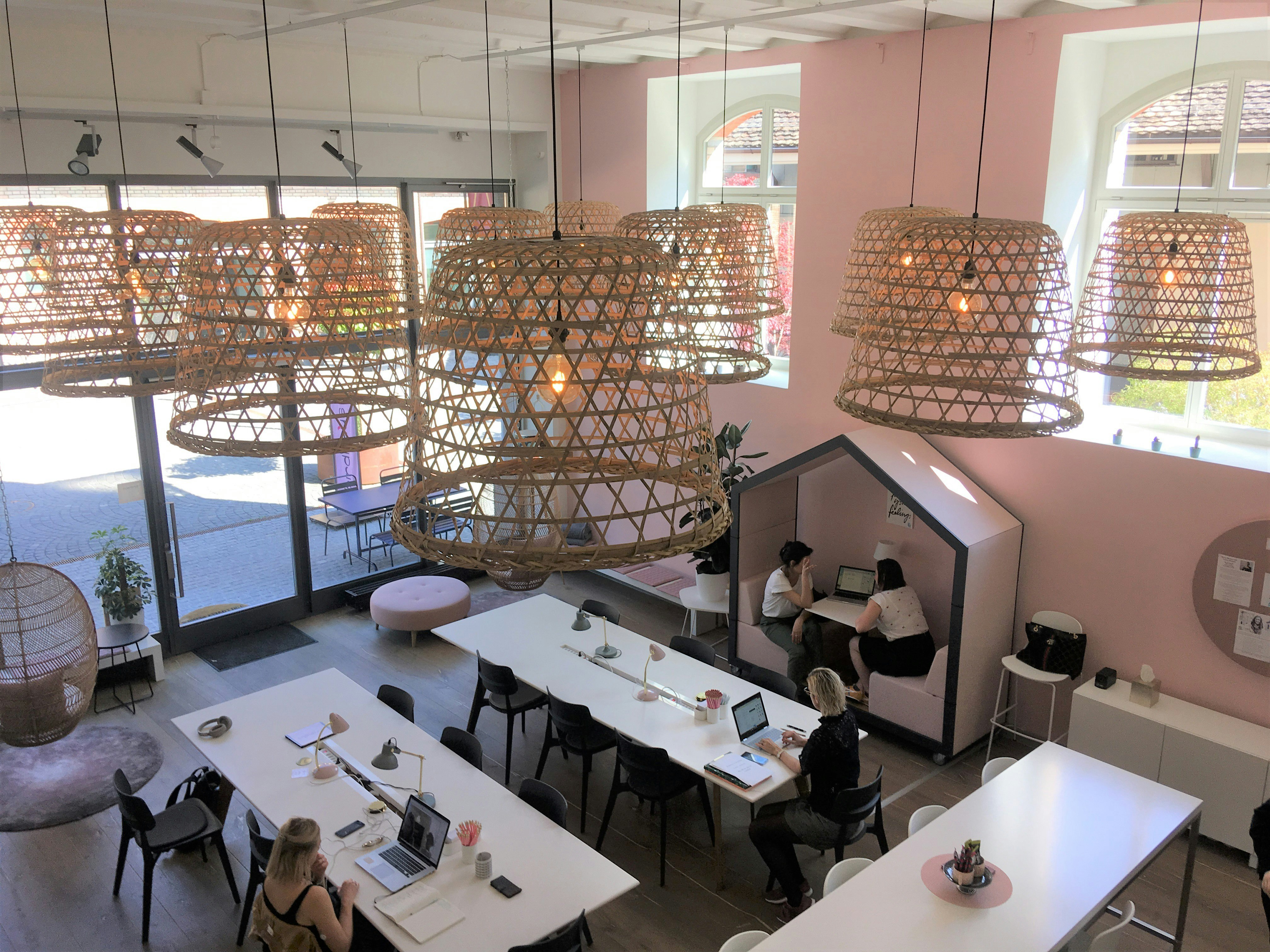 Looking down from a mezzanine floor to the high-ceilinged interior of Birdhaus: women are working at white tables, there is a pink wall with two high windows, and many oversized wicker lampshades overhead.