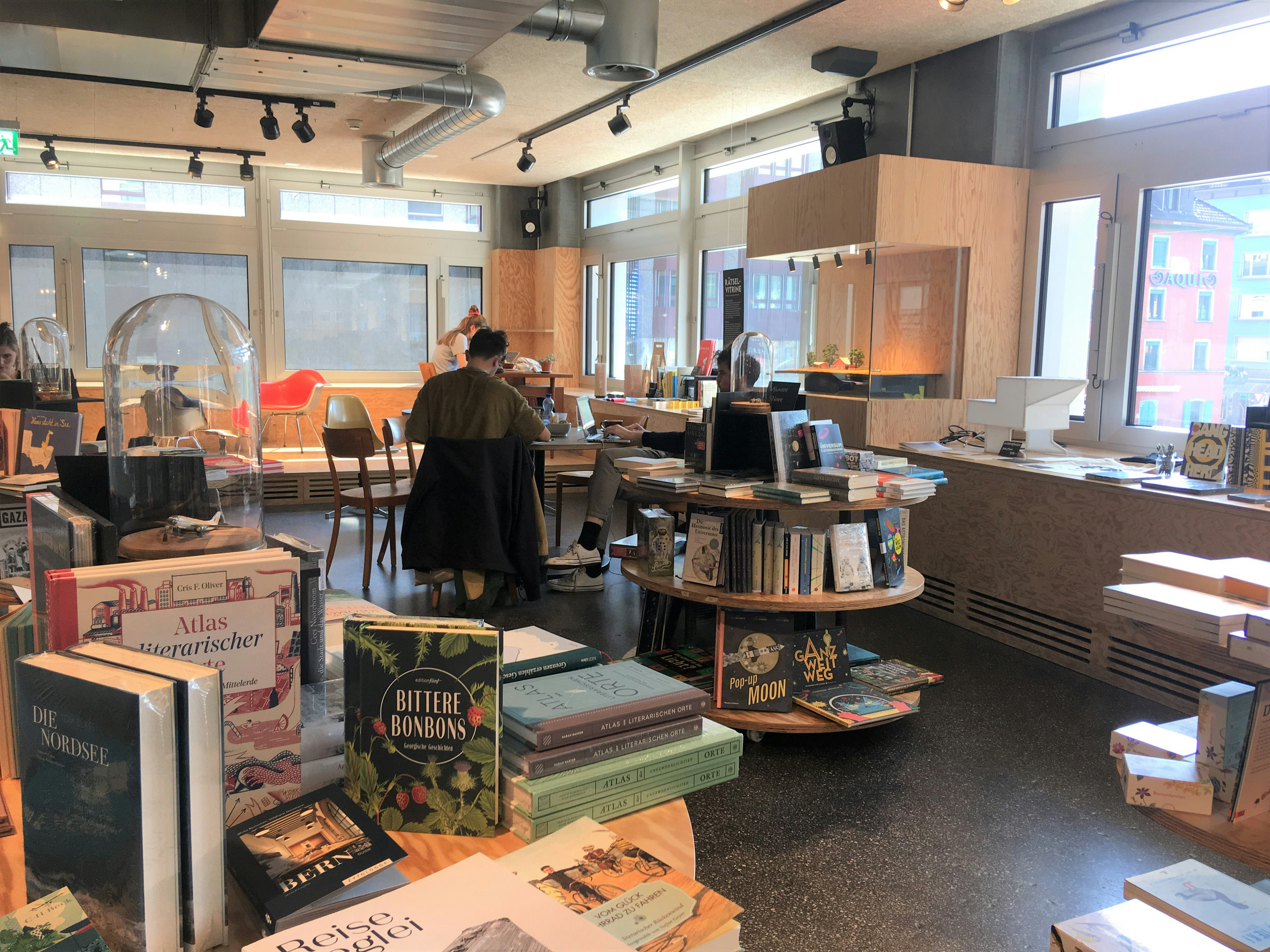 Circular display tables are covered with books for sale in Kosmos: beyond, customers are working at tables while the walls are lined with windows looking over other buildings outside. 