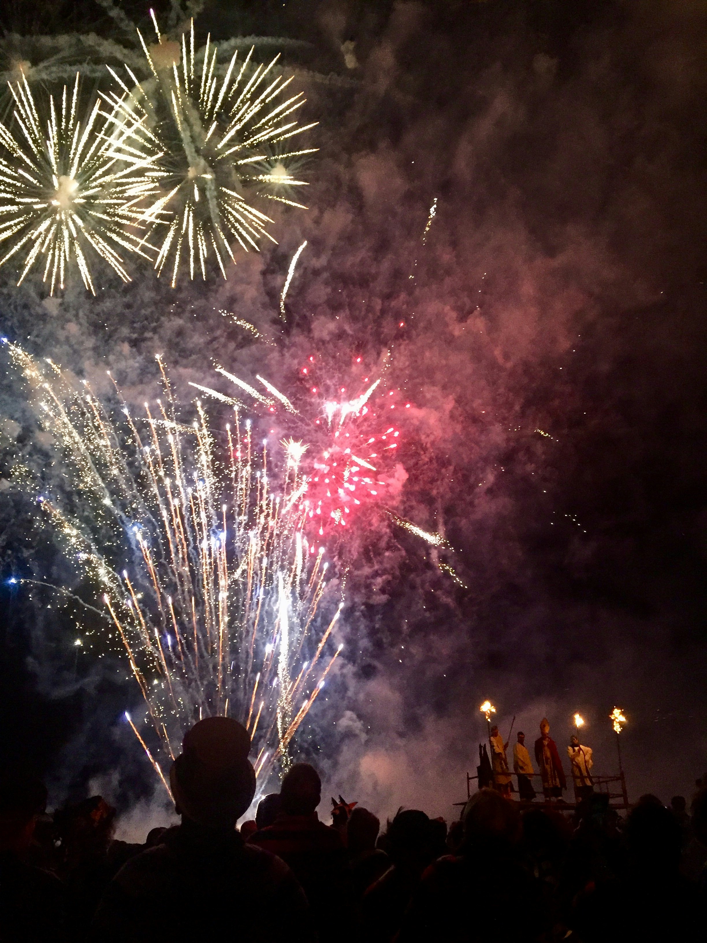 A nighttime scene of fireworks lighting a dark sky. The heads of people standing in the crowd are silhouetted against the bright fireworks.