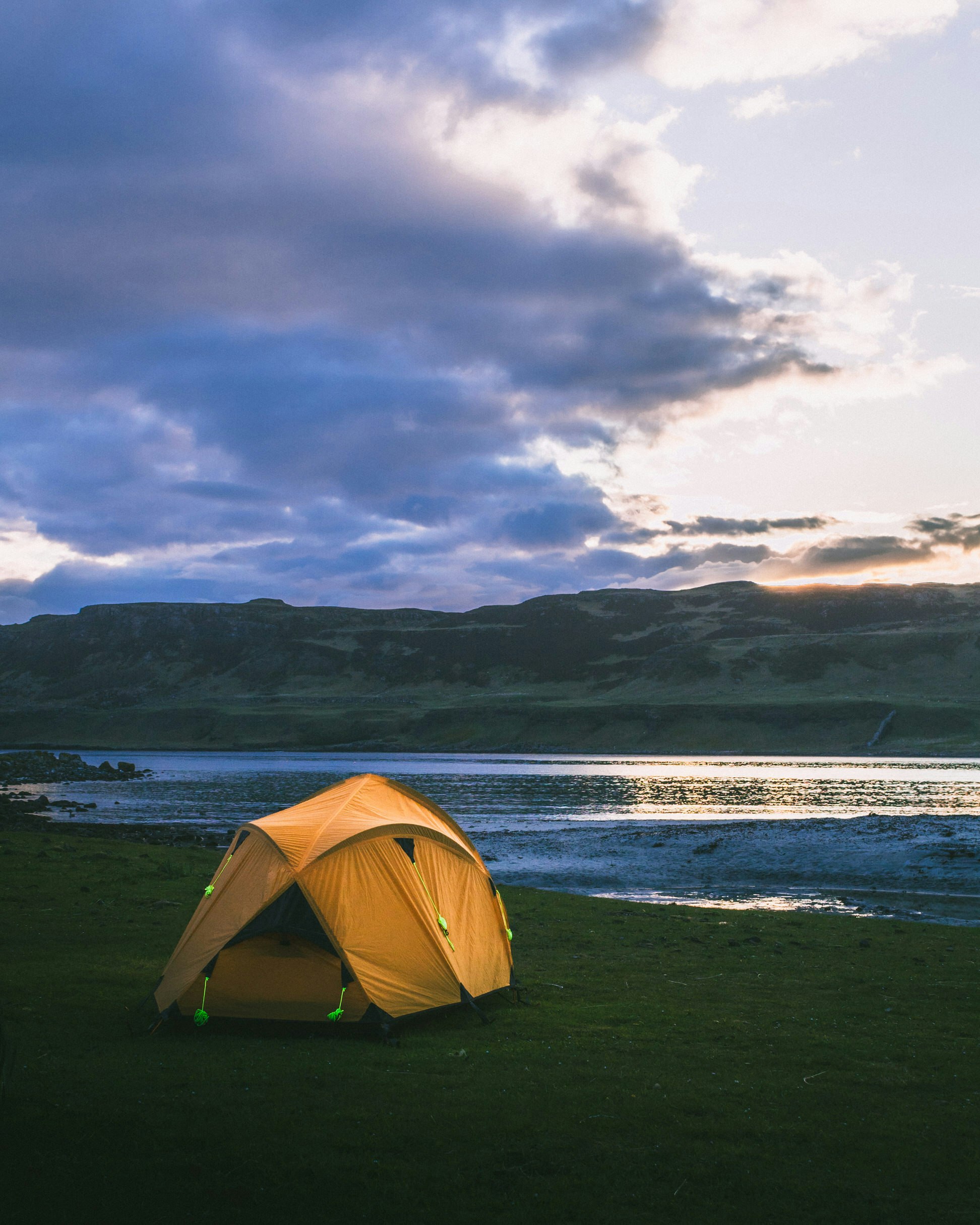  A bright yellow tent has been pitched on grassland just behind a shoreline. It's dusk, and the sun has just dipped below the hills in the distance.