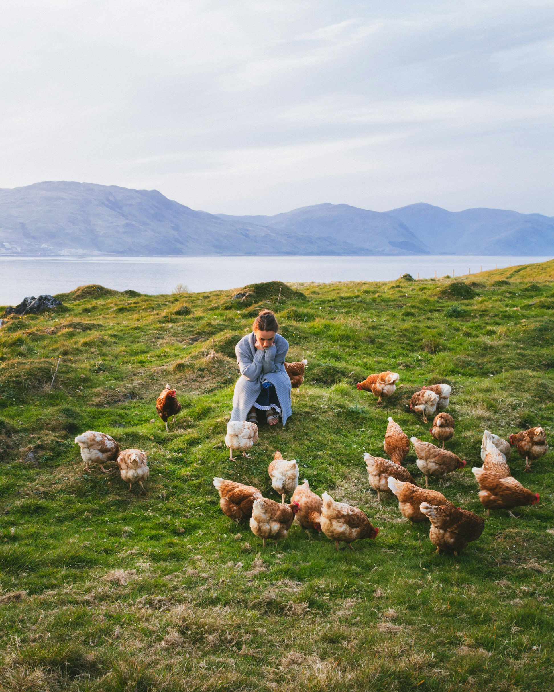 The writer is crouched down on grassy land amongst a flock of hens; there is a loch behind her and, beyond it, low mountains.