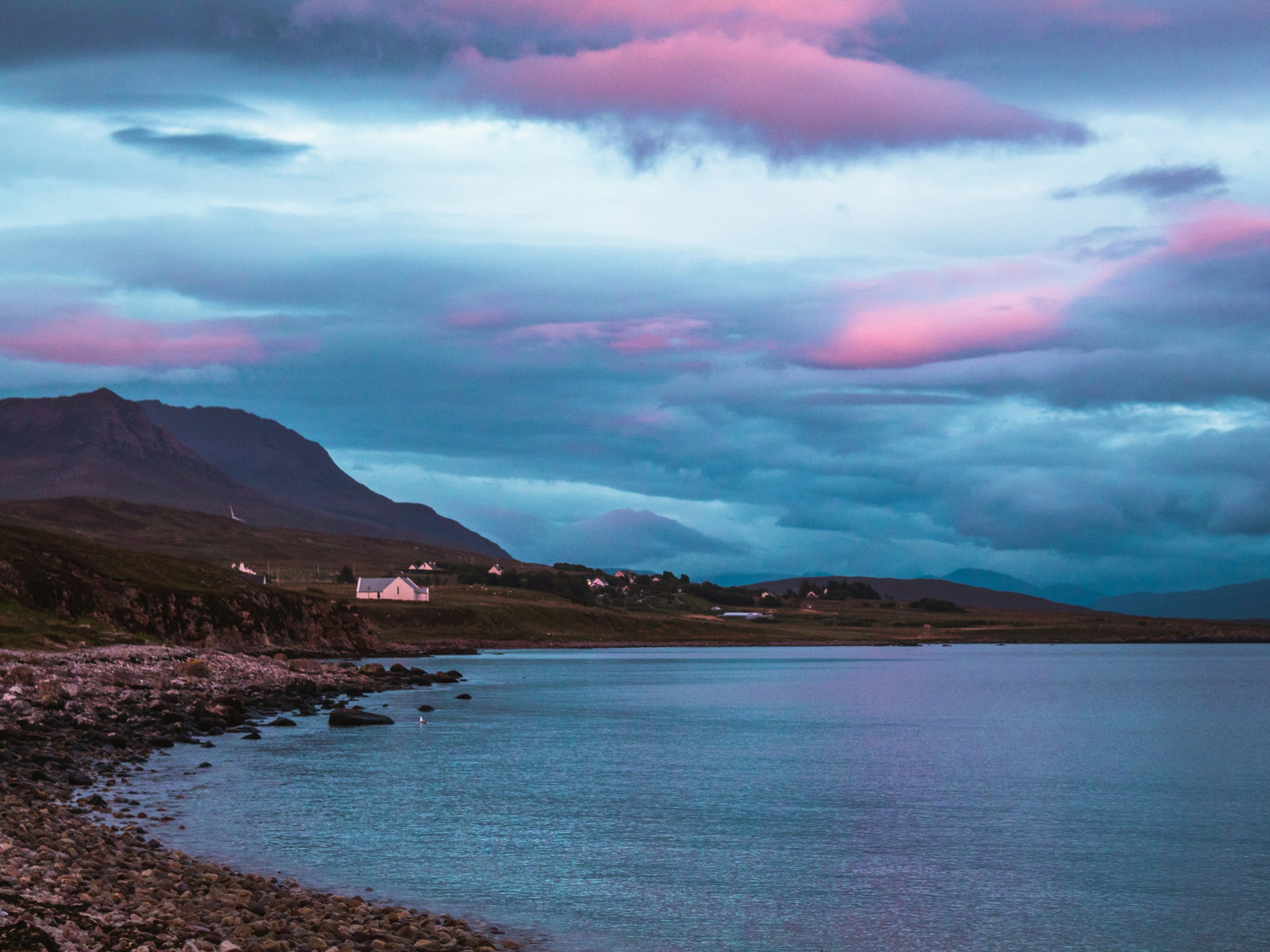 A pink and grey sky hangs above a hilly shroreline. In the distance is a hamlet of houses.