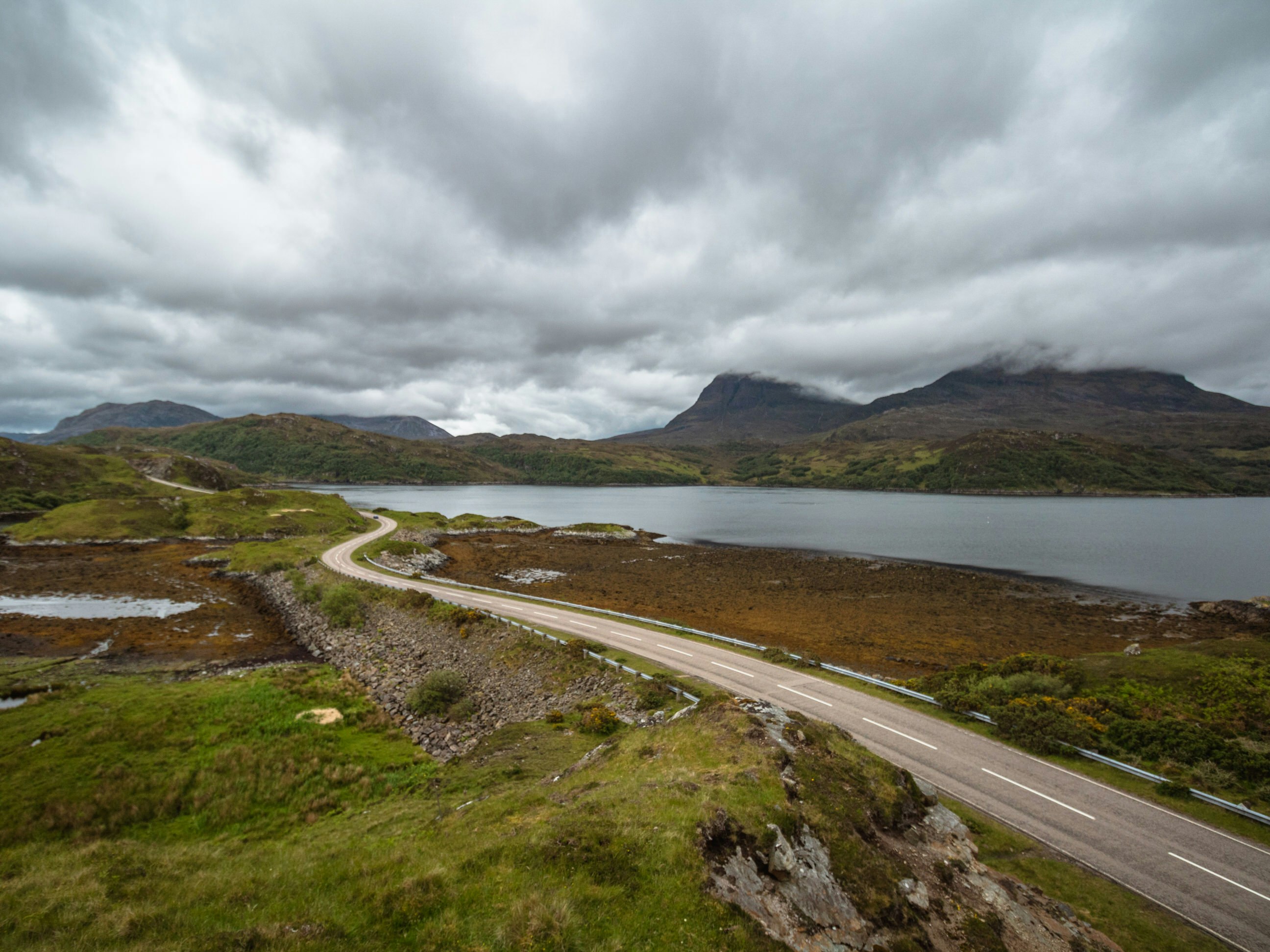 A narrow road winds a path beside a body of water in the Scottish Highlands; grey clouds linger on the low hills ahead.