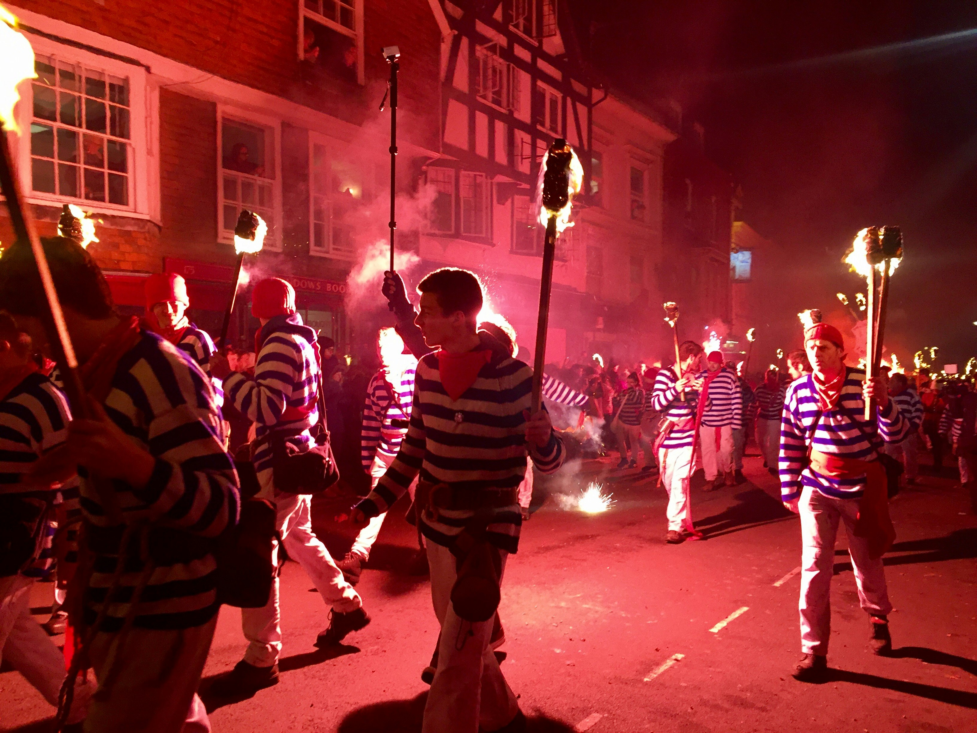 Scores of men and women in matching costumes – white trousers, blue and white striped t-shirts and red neckerchiefs – parade down a narrow street at night, holding flaming torches aloft.