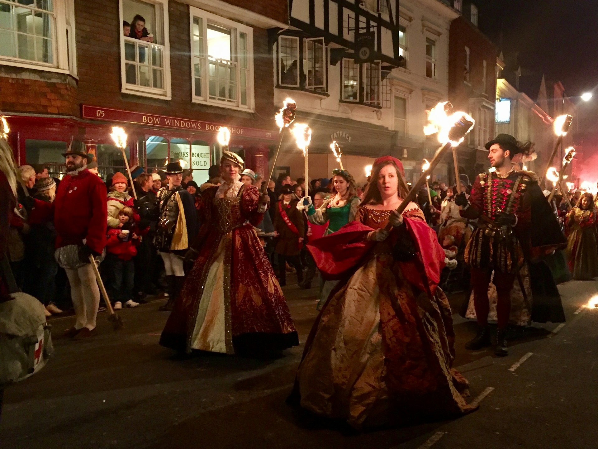 Men and women wearing elaborate historical costumes hold flaming torches as they parade down a street in Lewes.