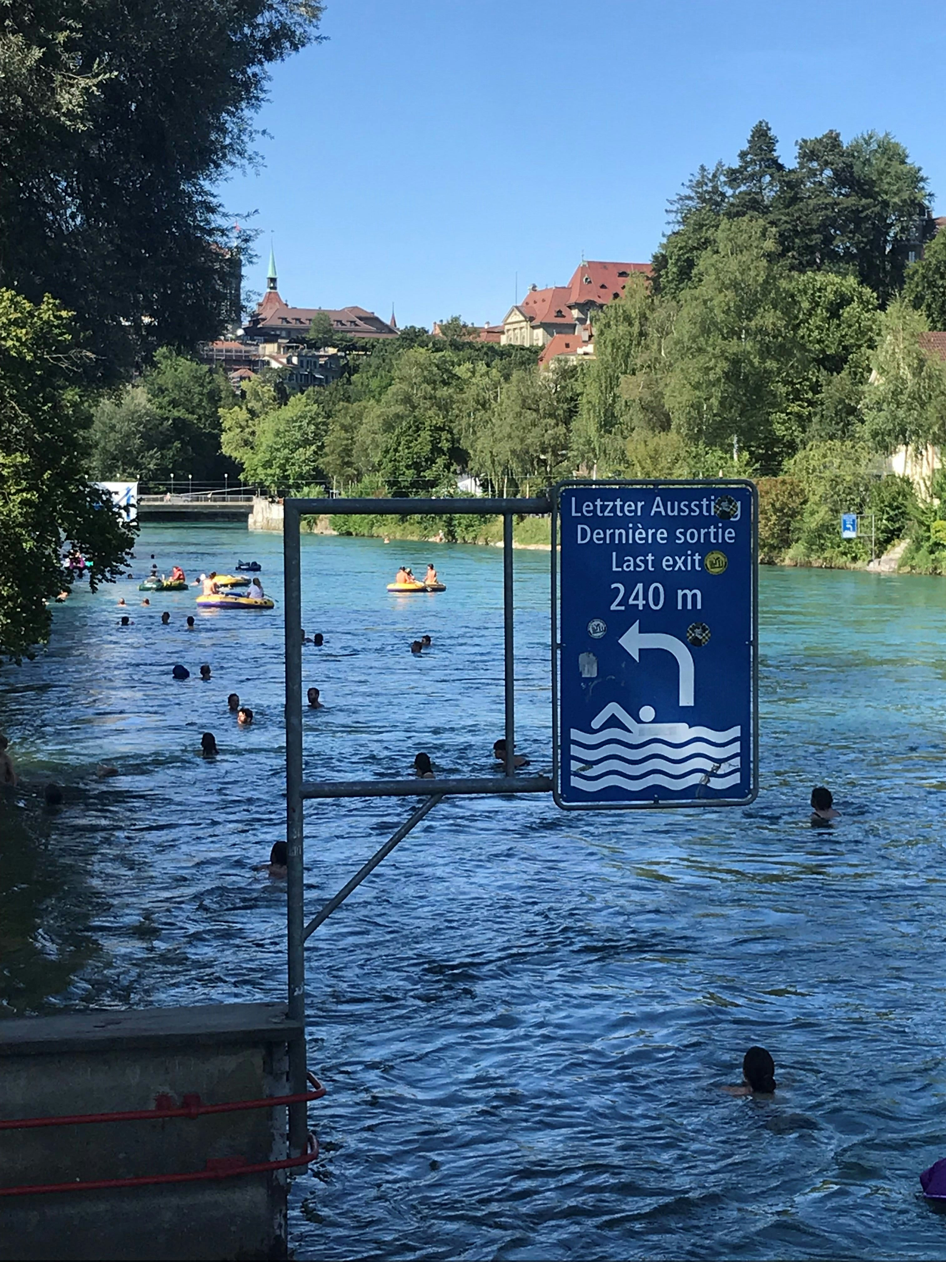 Many people are swimming down a river. Some groups are in yellow inflatable rafts. In the foreground is a large blue sign indicating the last exit for swimmers to leave the river.