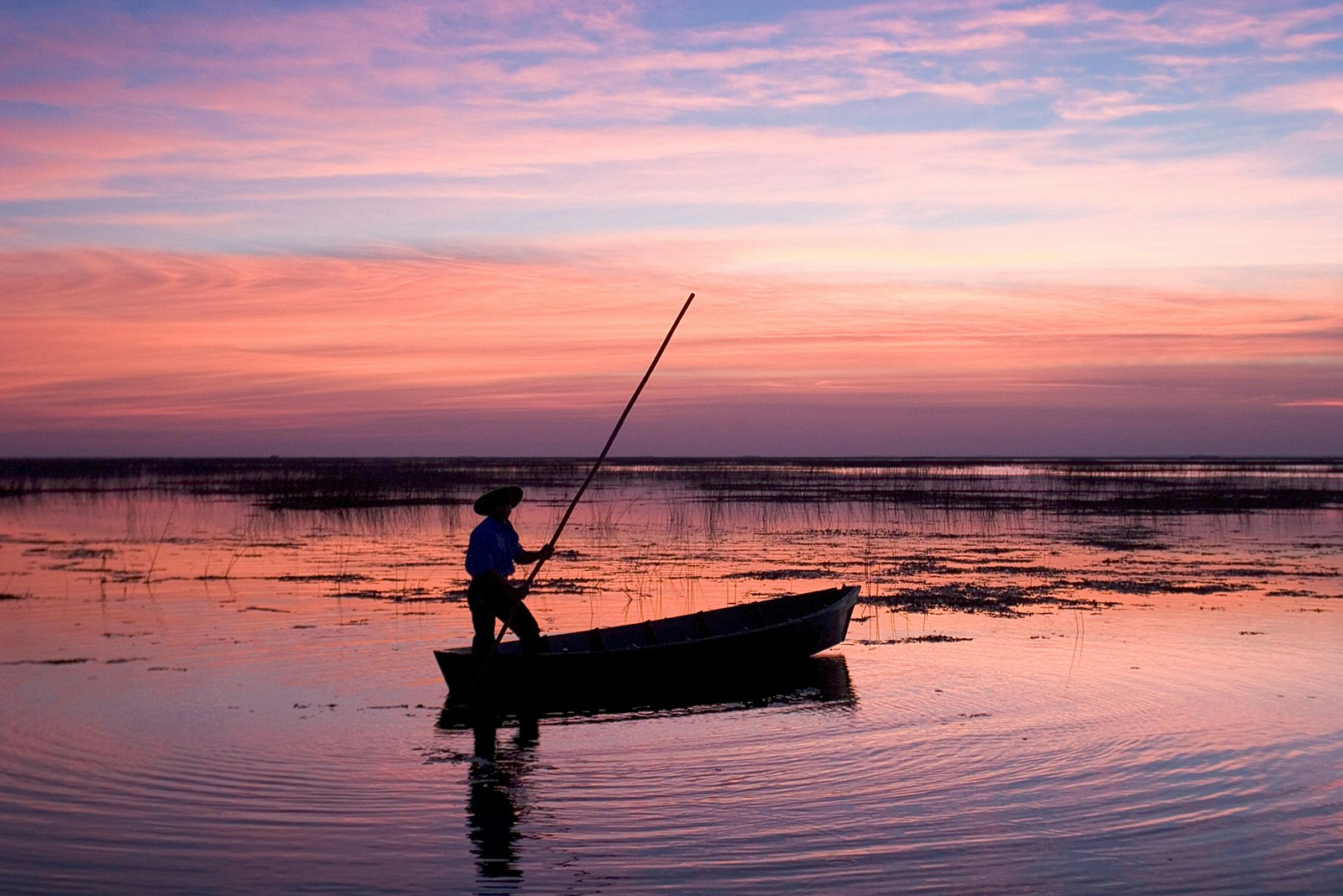 A silhouette of a person wearing a hat, standing in a boat holding a long pole at sunset. The wetlands reflect pink and purple. Iberá Wetlands, Northeast Argentina.