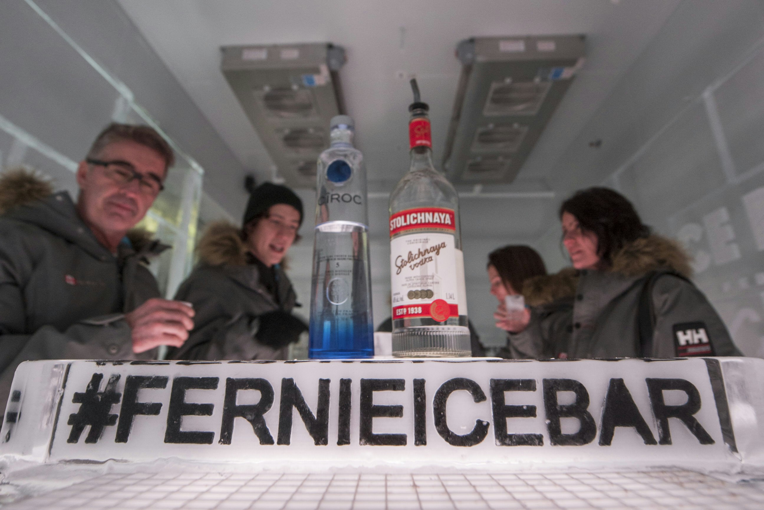Five people are seated around a table in an ice bar, chatting. One of the people is obscured by two bottles of alcohol in the foreground. In front of those bottles is a sign made of ice with the bar's official hashtag.
