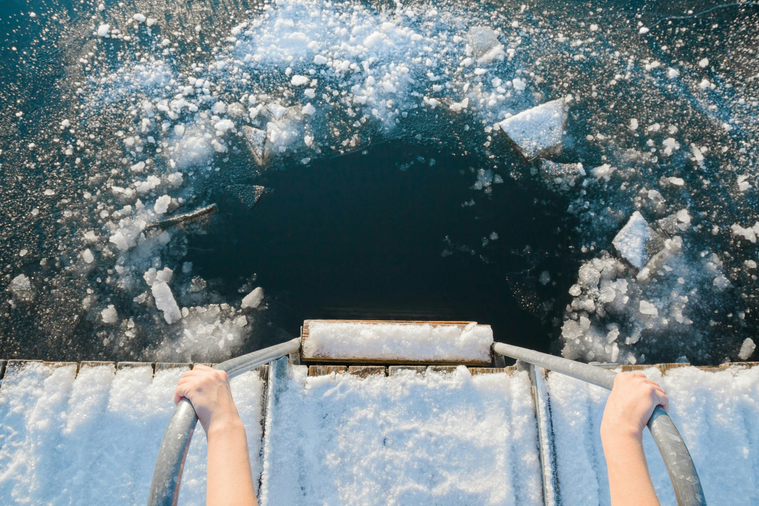 A point-of-view shot downwards into the frozen water. Hands hold on to railings and the shot focuses on the hole in the ice.
