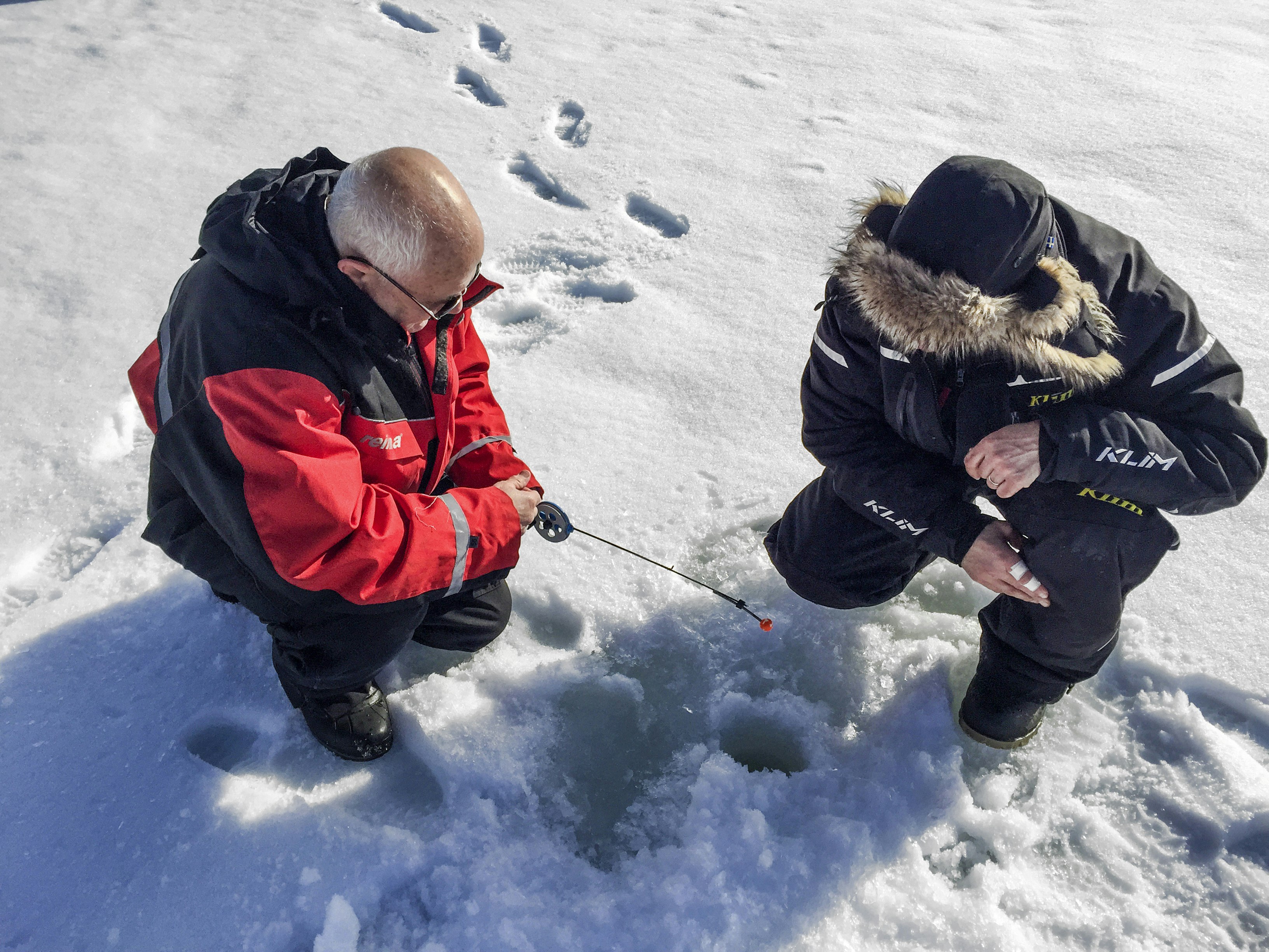 A pair of men sit in front of a hole drilled into the ice for ice fishing 