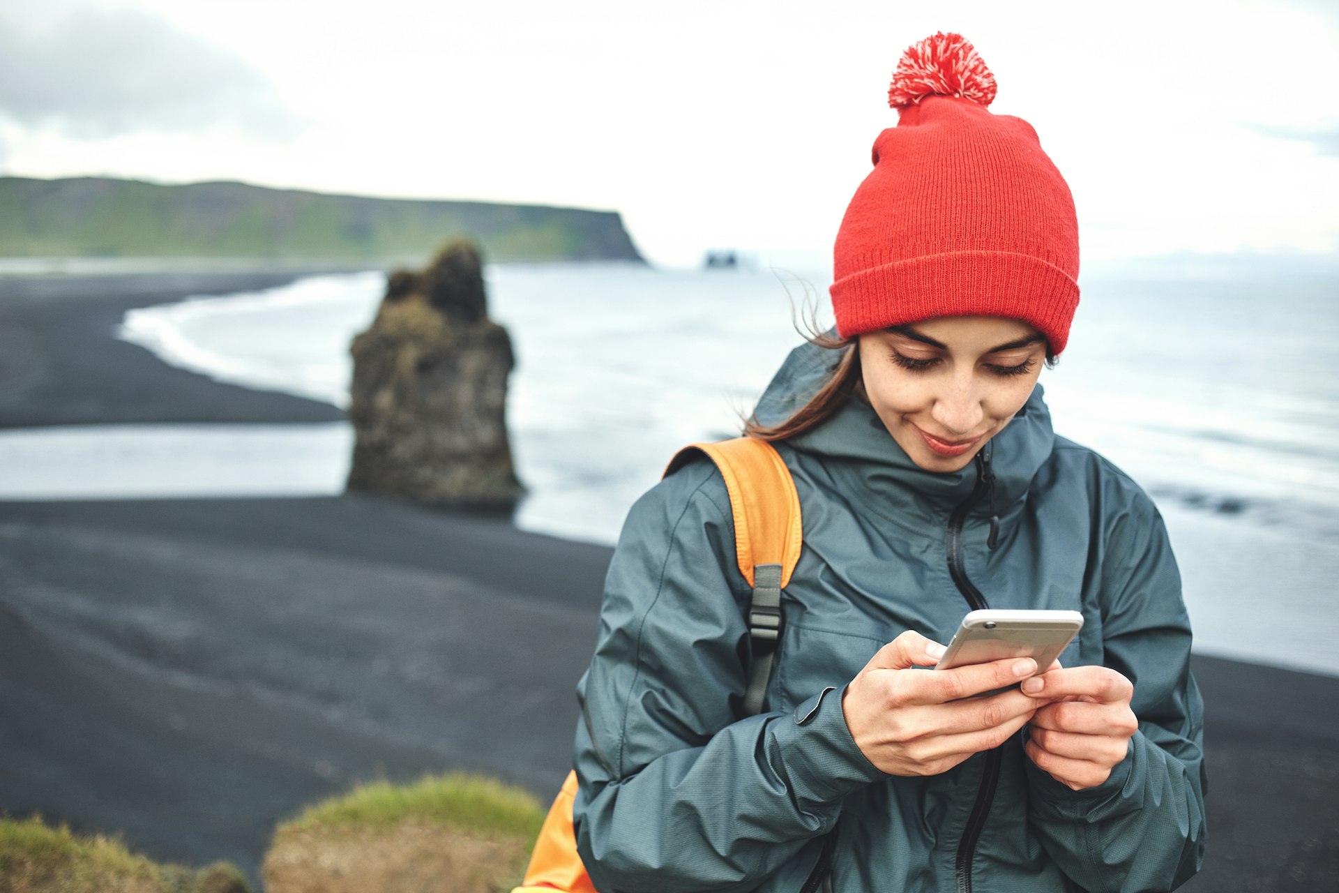 A woman wearing a bright red hat uses her phone on a black beach in Iceland. 