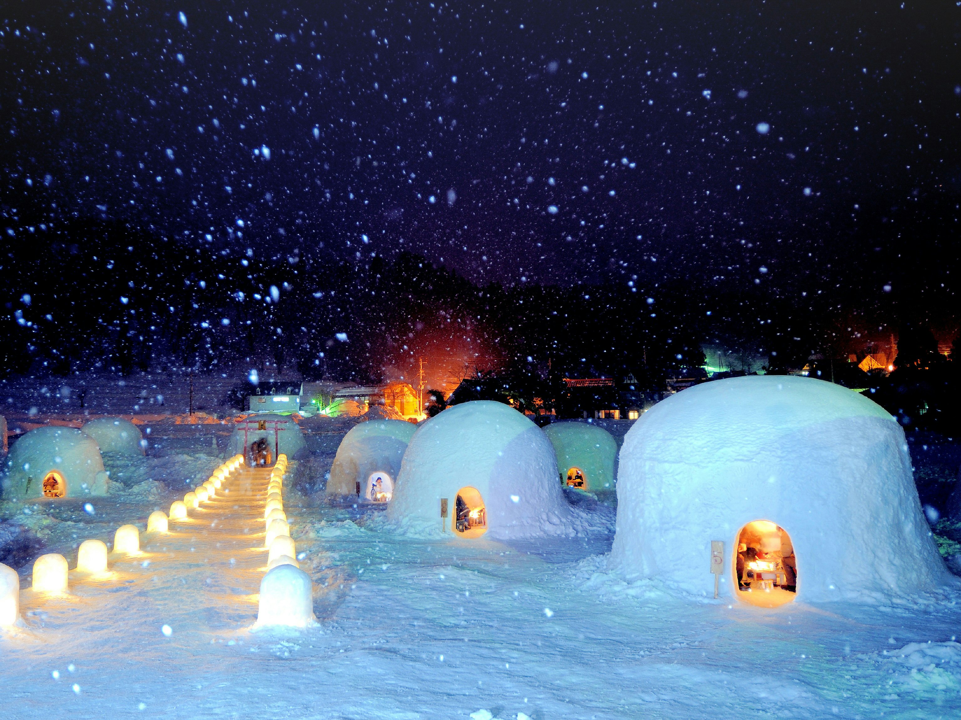 A snowy, night-time scene of a collection of igloos at Iiyama Kamakura Village in Nagano; through the igloos' small arched doors, people are visible in the latern-lit interiors. 