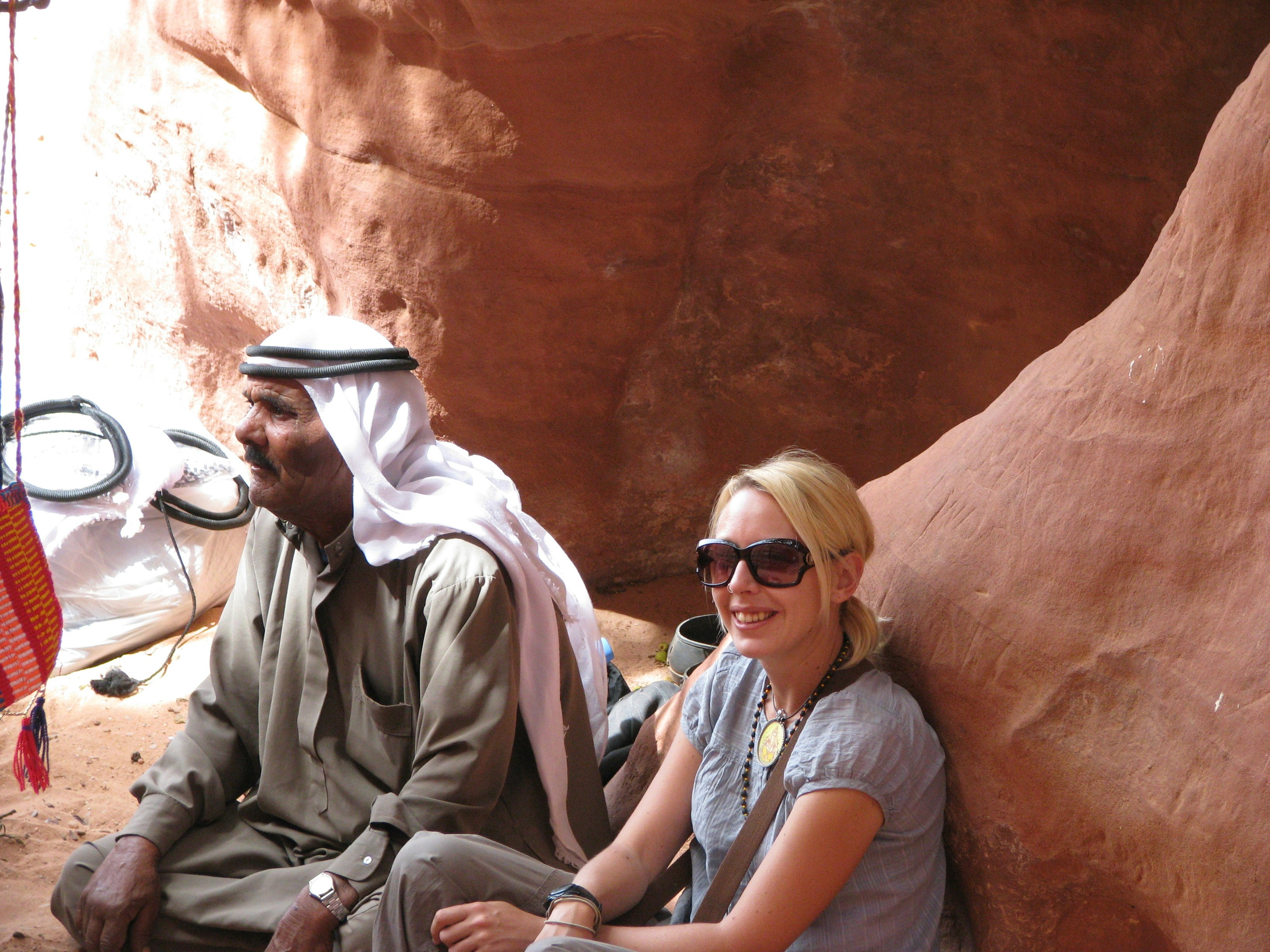 Jess sitting at the foot of a red cliff at Wadi Rum, Jordan; next to her is a local wearing a ghutrah (traditional Arab headdress).